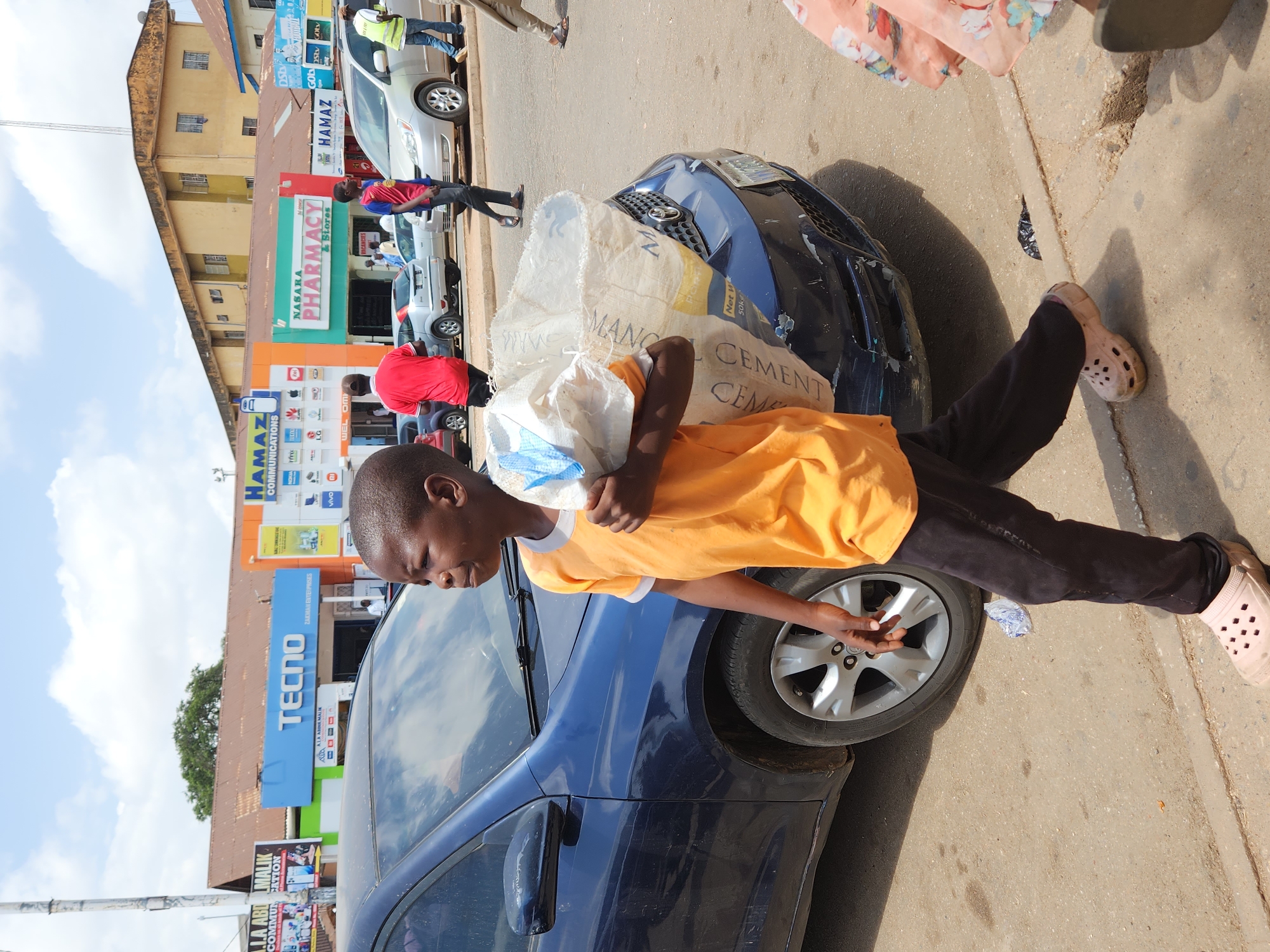 A young person carries a large bag of cement across the hood of a dark blue car on a street. The person is wearing an orange shirt, dark pants, and pink shoes. The car is parked on the right side of the street. Behind the car, there are shops with signs for Tecno, Nasaka Pharmacy, and Hamaz Communications. There are also other cars and people visible in the background. The sky is blue with some clouds.