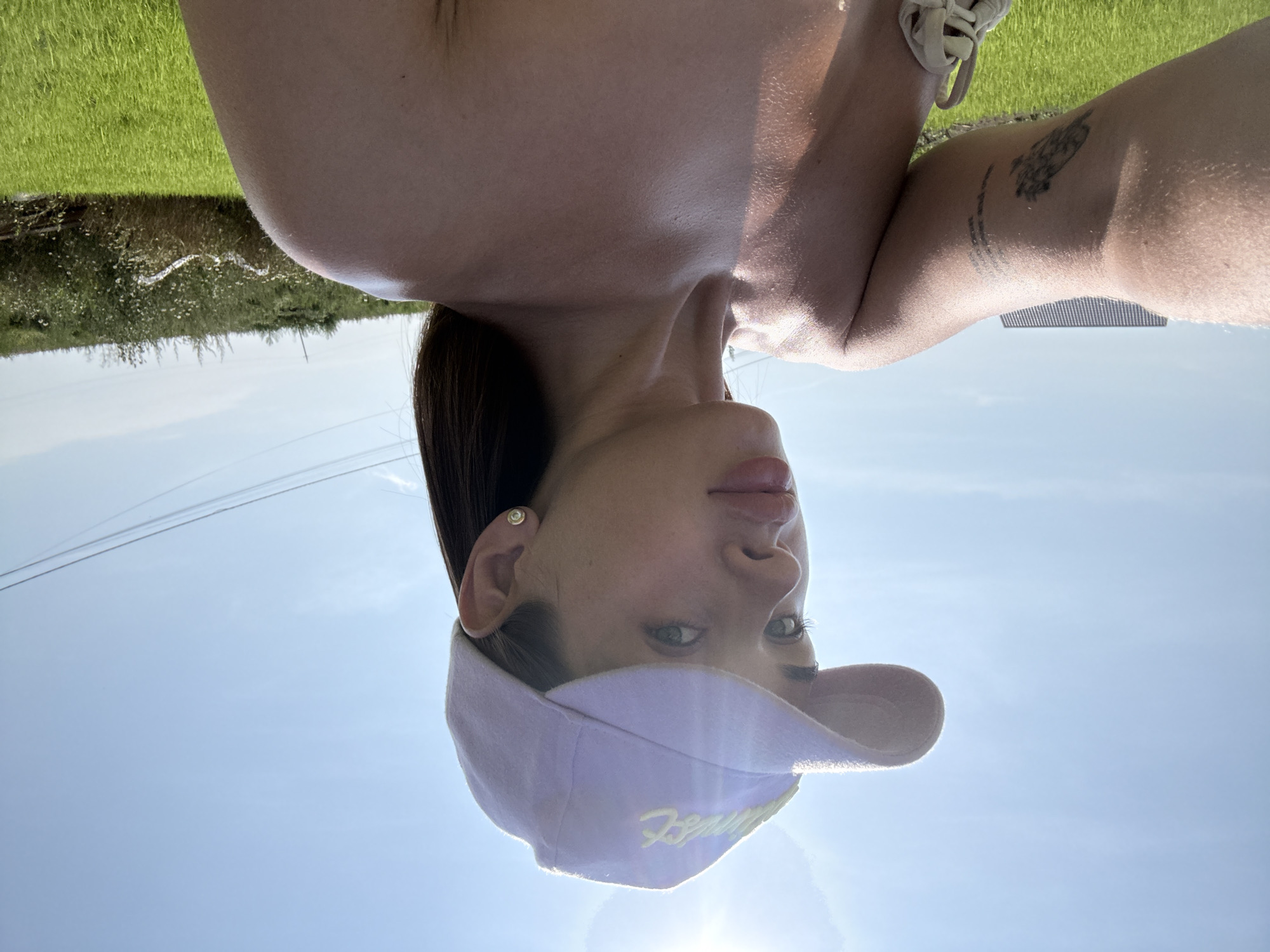 A young woman is captured in a close-up, upside-down selfie taken outdoors. She is wearing a light purple baseball cap with yellow embroidery that reads "Fenty." She has dark hair, and her face is partially obscured by the cap. A pearl stud earring is visible in her left ear. Her skin appears sun-kissed, and there's a hint of a tattoo on her right arm.
The background suggests a sunny day. To the upper left, a blurred image of green grass and some foliage can be seen, indicating a rural or park-like setting. The rest of the background is a bright, clear blue sky, with a hint of a sunflare. The orientation of the image implies the photo was taken outdoors during the daytime, likely in the afternoon given the sunlight. The overall mood appears casual and relaxed. No specific activity is discernable beyond the act of taking a selfie.
The location context is Лобаново, Russia, though there are no overt visual cues in the image to confirm this specific location.