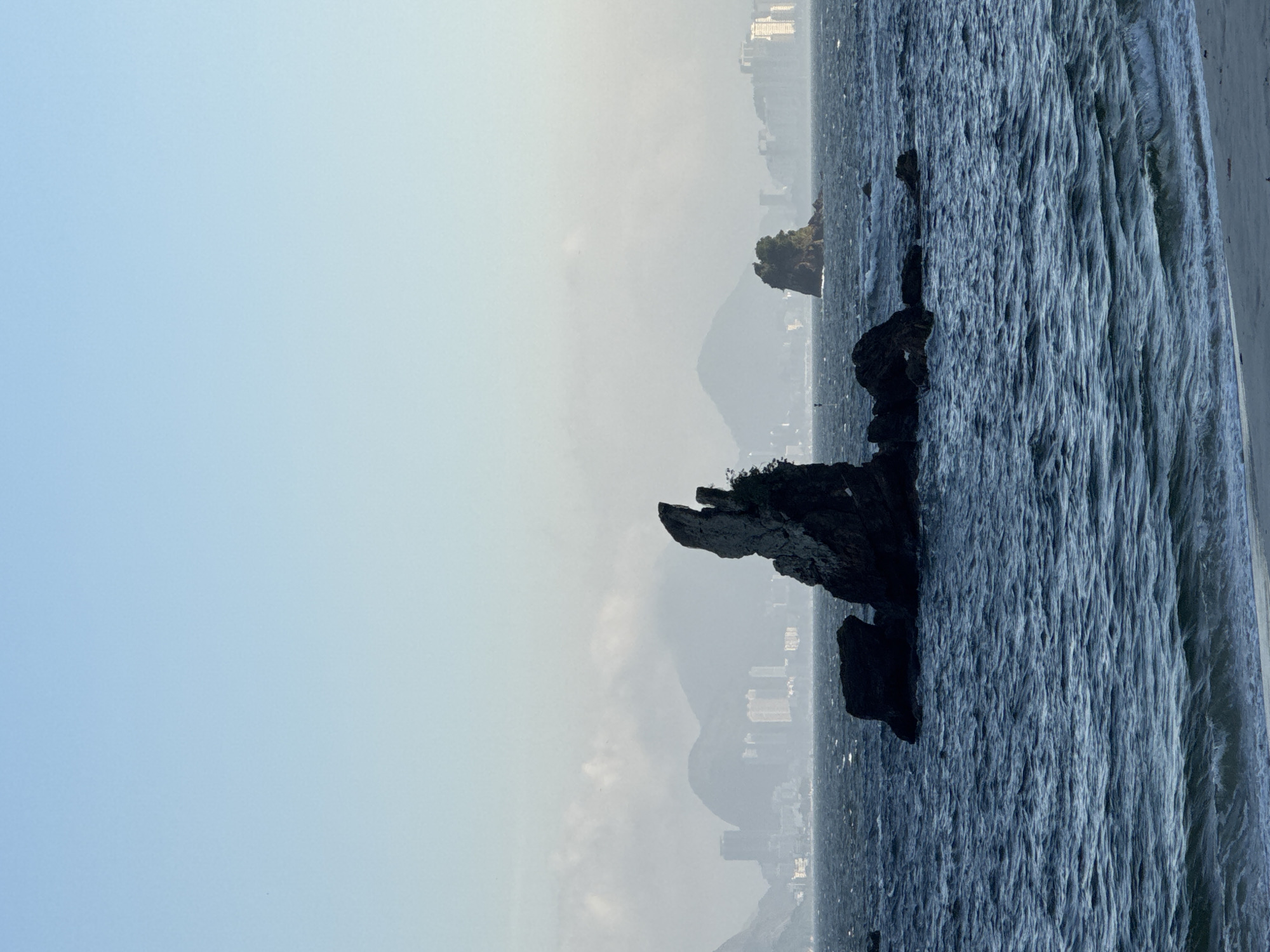This is a landscape photograph taken from the shore of the city of Niterói, Brazil. The image features a dark, jagged rock formation in the foreground, rising prominently from the choppy, dark blue sea. To the right and further back, another smaller, tree-covered rock formation is visible. In the background, the cityscape of Niterói, with its buildings and mountains, is shrouded in a light haze under a pale blue sky. The sea exhibits small waves and ripples, indicating a moderate breeze. There are no people or animals present in the scene. The overall mood is serene and natural, highlighting the interplay between the urban environment and the coastal landscape.