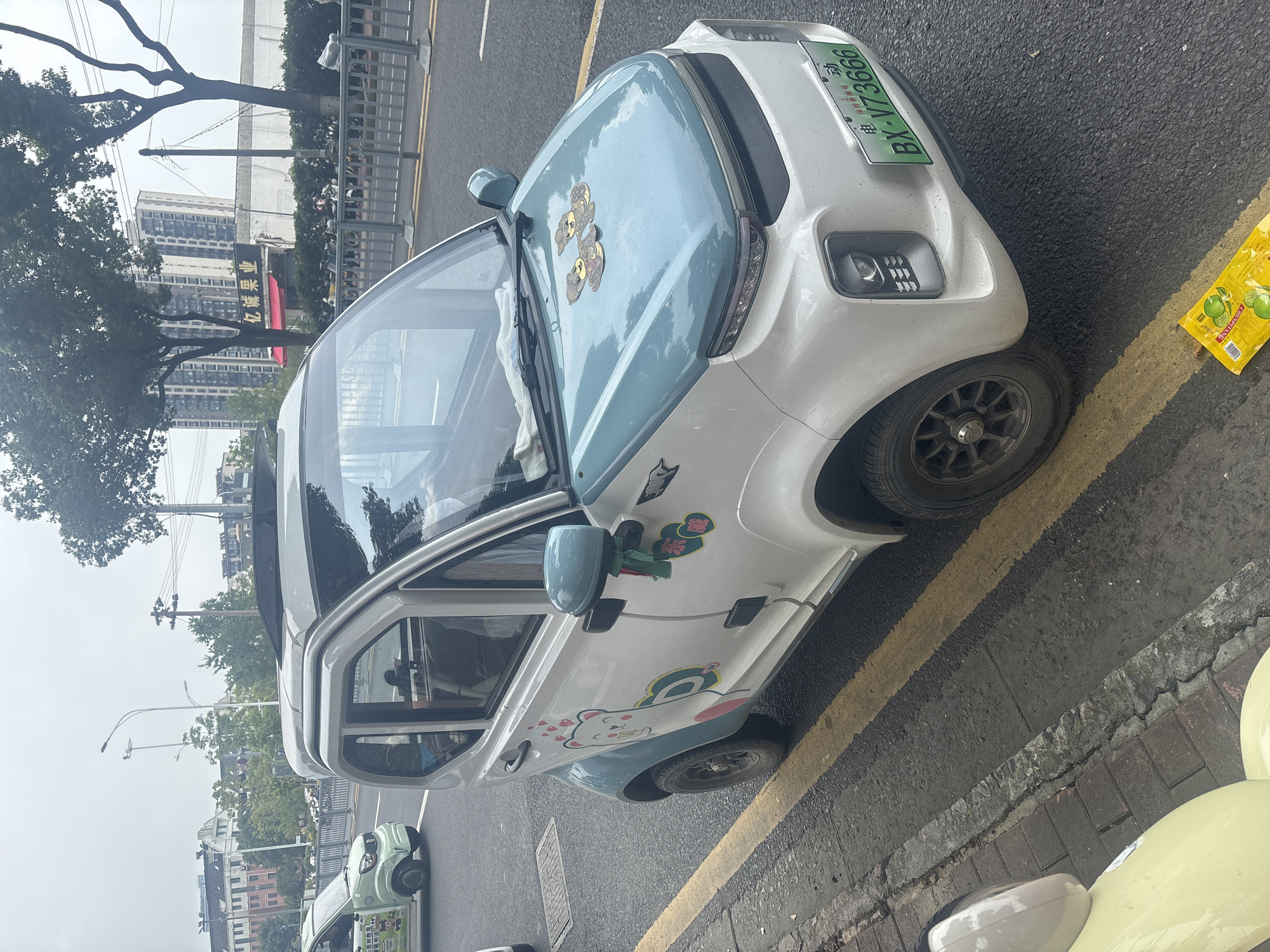 A white and light blue electric mini-car is parked on the side of a street in Suzhou Industrial Park, China. The car is decorated with cartoon stickers of a bear and other characters. It has a license plate with the characters "BX-V73666". The street has yellow double lines. In the background, there are buildings, trees, and streetlights. A light green mini-car is visible in the distance. A yellow package is on the ground near the car.
