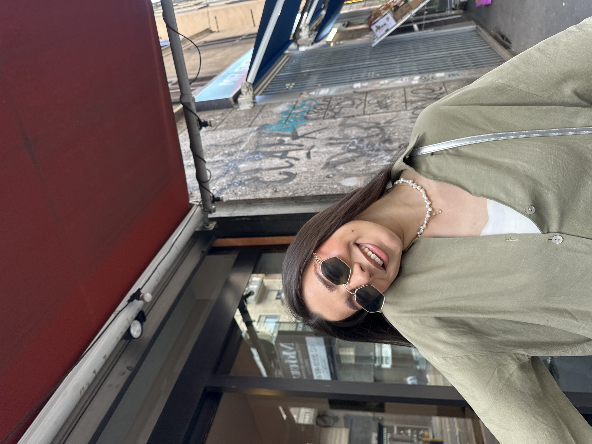 The image shows a woman standing on a street in Milan, Italy. She is wearing sunglasses, a pearl necklace, and a light green shirt or jacket. Behind her is a building with a red awning and a storefront. The street is paved with stone and has graffiti on the walls. A blue awning is visible in the background. The buildings are reflected in the storefront's glass.