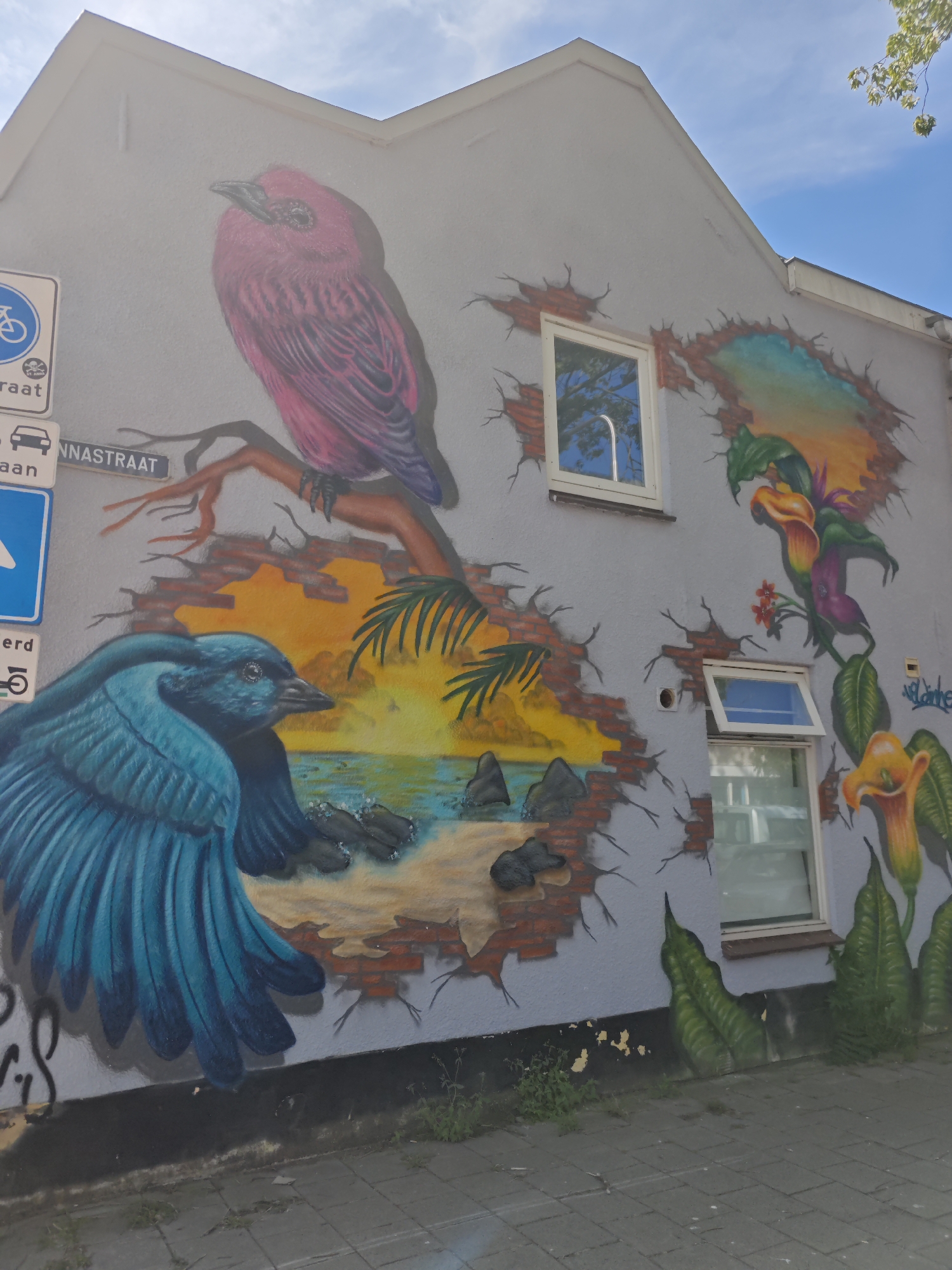 The image shows a mural on the side of a building located on Annastraat in Tilburg, Netherlands. The mural depicts two birds and a tropical landscape. On the left side of the mural, a large pink bird is perched on a branch, and below it, a blue bird with its wings spread is in the foreground. A scenic view of a beach with palm trees and a sunset can be seen through a brick wall that appears to be broken. On the right side, there are large calla lily flowers and lush green foliage. The building has two windows visible in the mural area, one of which is open. Traffic signs are present on the left side of the building. The ground in front of the building is paved with gray bricks. The sky is blue with some clouds.