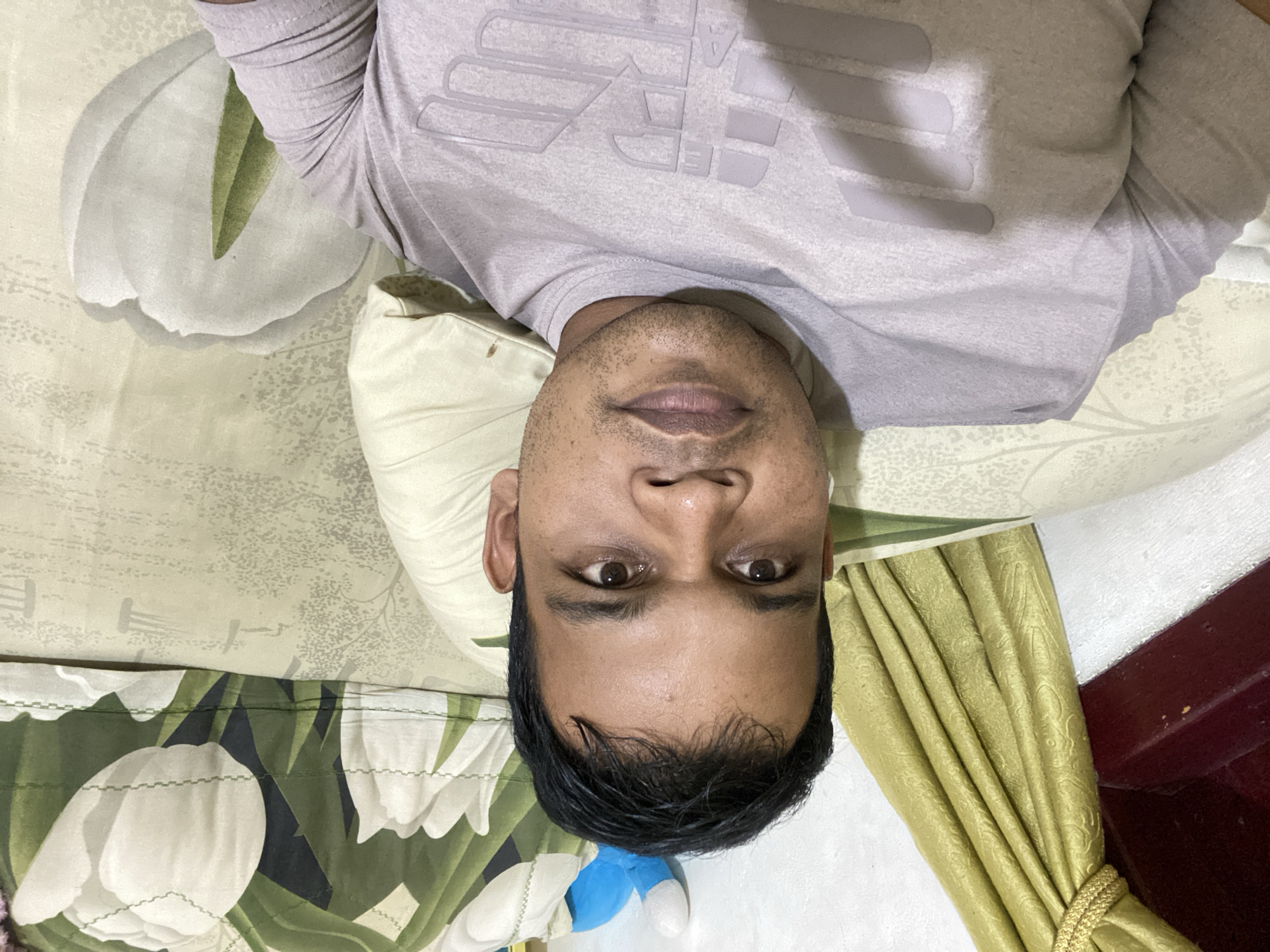 A medium shot, chest-up, selfie style photo shows a South Asian man with dark hair and brown eyes lying down. He is wearing a light purple t-shirt with a large, light gray eagle logo on the chest. The man is on a bed with a cream colored duvet that has a pattern of large white tulips and green leaves. To the left of the man is a golden curtain. Behind him, and slightly to the right, is a pillow, and a blue stuffed toy is visible above the pillow.