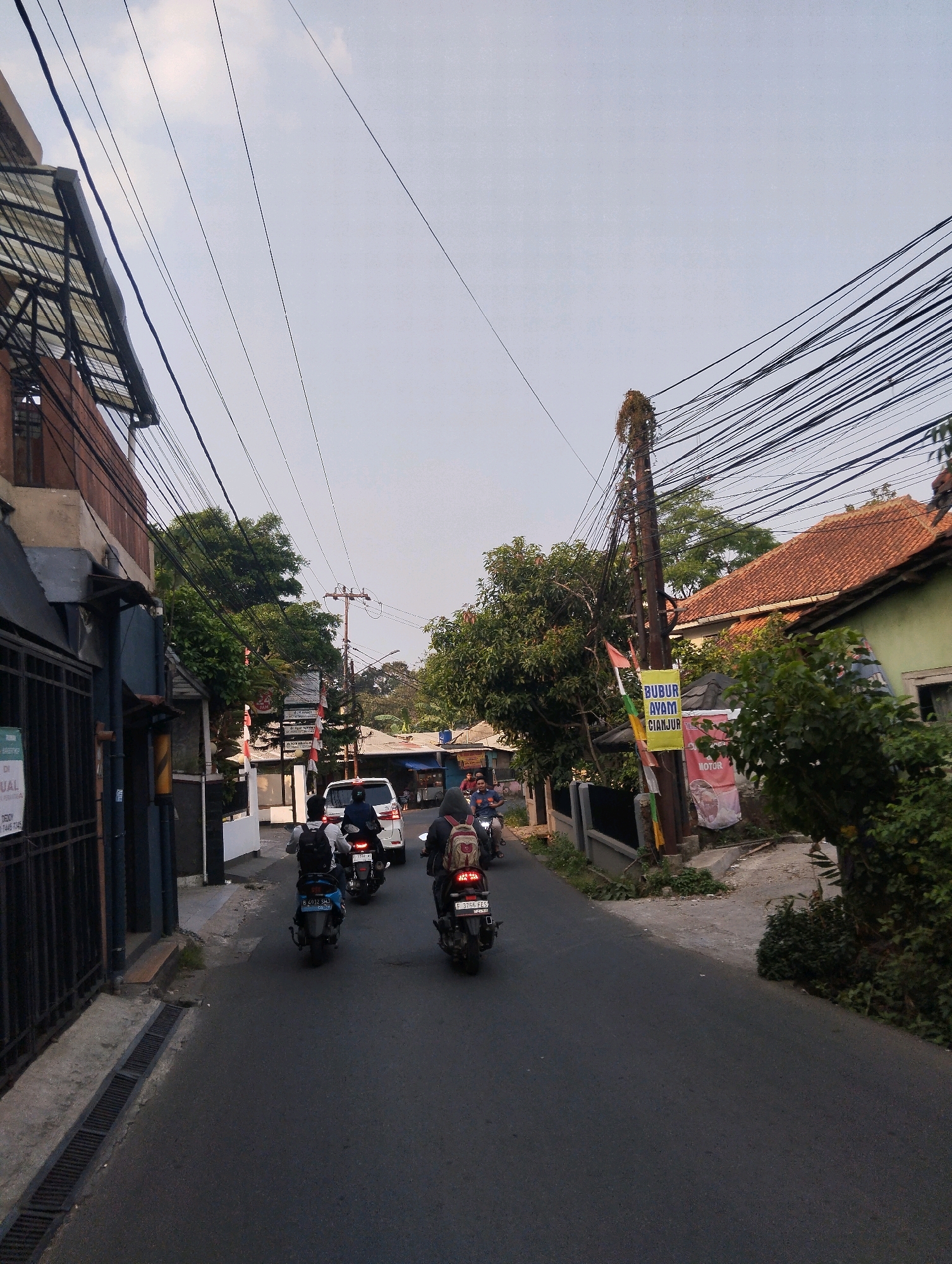 The image depicts a street scene in Sukaraja, Indonesia, on a cloudy day. Several motorcycles are traveling down a paved road, with riders wearing backpacks. A white car is also visible on the road. On the left side of the street, there is a building with a metal fence and some windows. On the right side, there are houses with red tile roofs, lush green trees, and a utility pole with numerous electrical wires. A banner on the utility pole reads "BUBUR AYAM CIANJUR". The overall atmosphere suggests a typical urban environment in Indonesia with active street life.