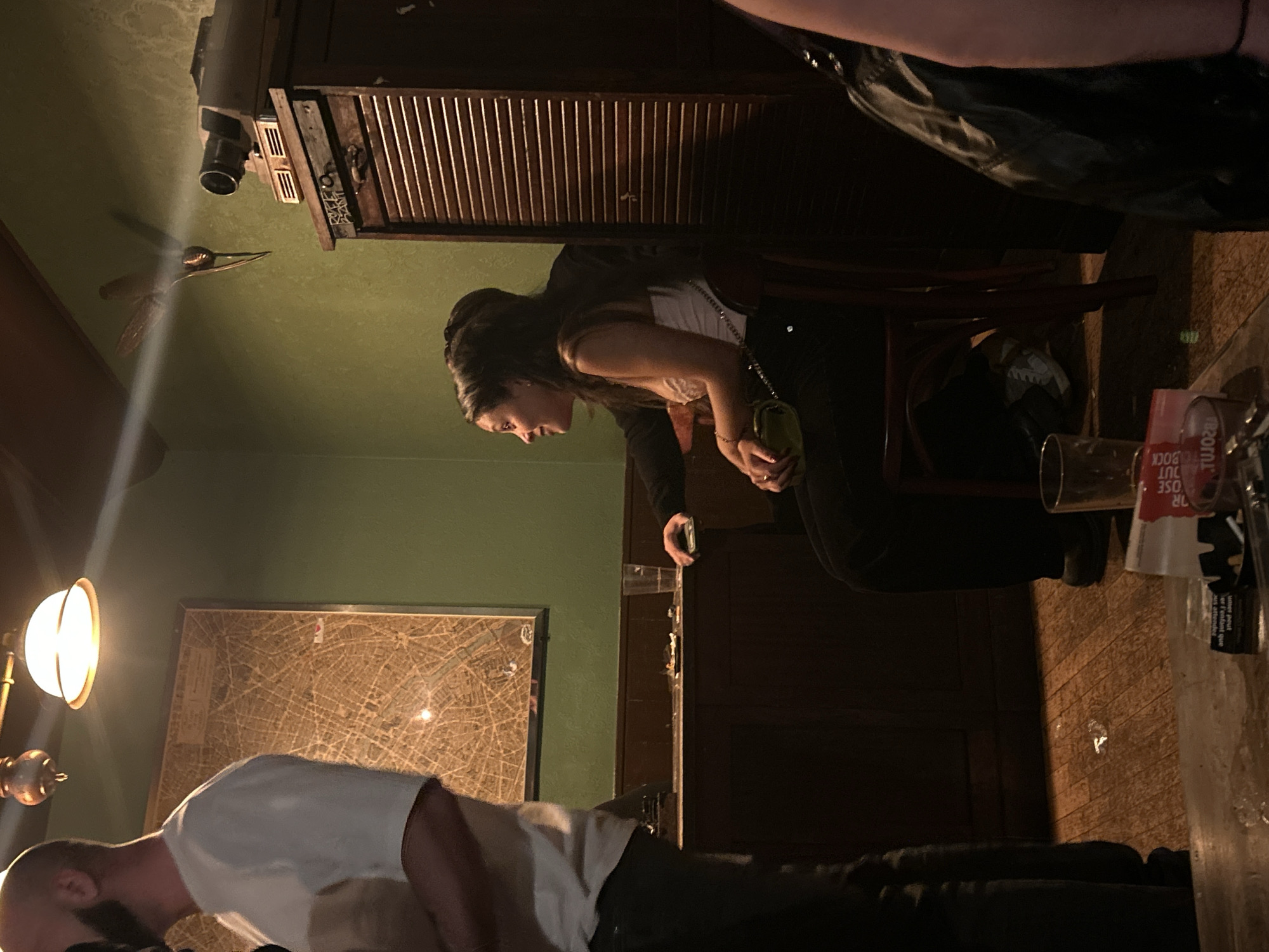 The photo depicts an interior scene, likely a bar, with several individuals present. A woman stands near a dark wooden structure, possibly a bar counter or cabinet, holding a glass in her right hand, with a small purse or bag hanging on her left shoulder. She's looking downwards towards a man.
The man is bent over or lying down, wearing a white t-shirt. A floor lamp is positioned near him, providing illumination. Behind him, there's a framed map of Paris, France on the wall. On the table to the right, one can see glasses and beverage-related items, suggesting an area for consumption.
The environment is dimly lit, creating a casual, relaxed atmosphere. The walls are painted in a muted green color.
