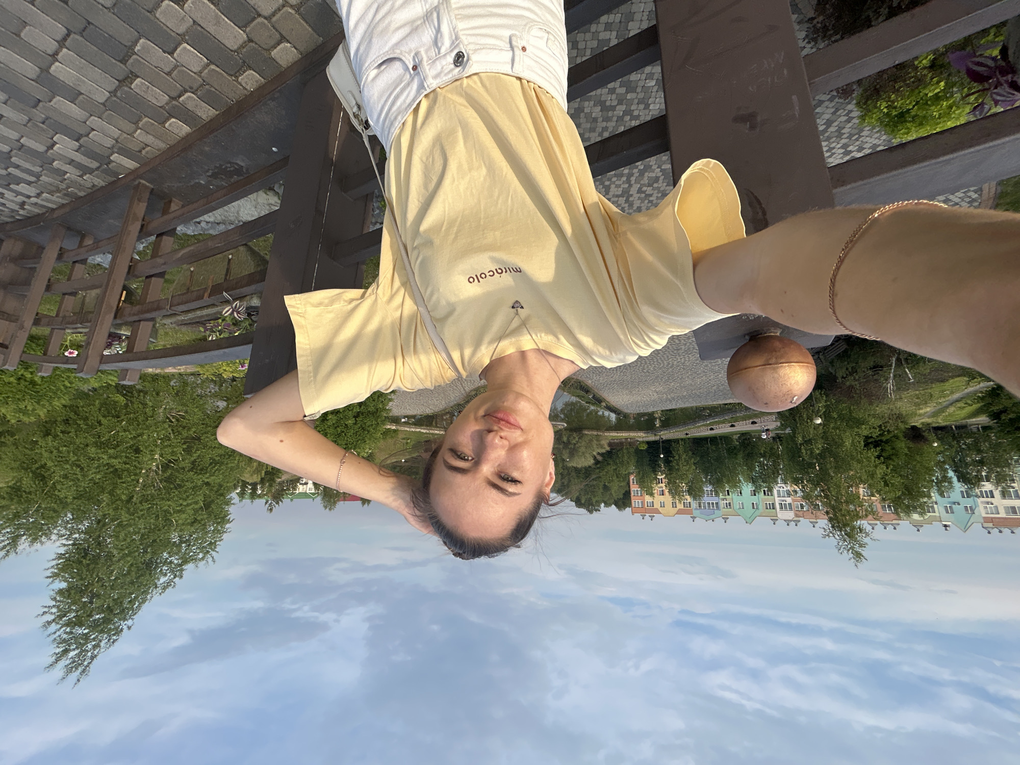 A young woman with dark hair pulled back and freckles across her nose takes a selfie outdoors. She is wearing a light yellow t-shirt with the word "miracolo" printed on it in brown lettering, and white denim shorts. A thin gold chain necklace with a heart-shaped pendant rests on her chest. Her left arm is extended, holding the camera, and her right hand is touching her head. She stands on a brown wooden bridge with a stone pathway. In the background, a row of colorful, multi-story apartment buildings line the opposite side of a calm body of water, surrounded by lush green trees. The sky is partly cloudy with a soft blue hue.