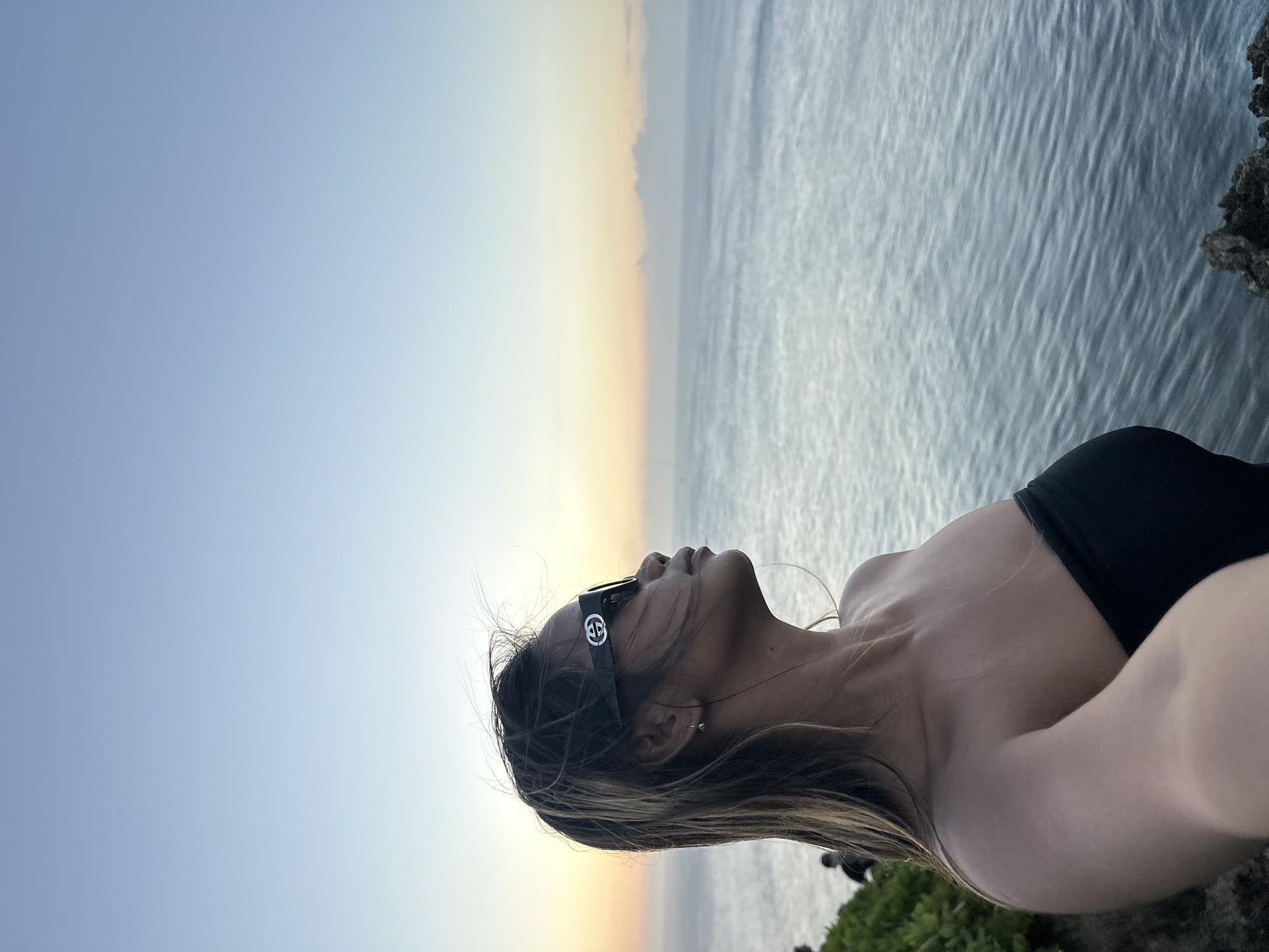 A woman wearing a black strapless top and sunglasses poses for a selfie on a cliff overlooking the ocean at sunset in Kuta Selatan, Indonesia. The sky is a gradient of light blue to pale yellow. The ocean is a deep blue, with small waves. The woman's hair is blowing in the wind. In the background, there are some green bushes and two people standing on the cliff. The woman is looking off to the side with a serene expression.