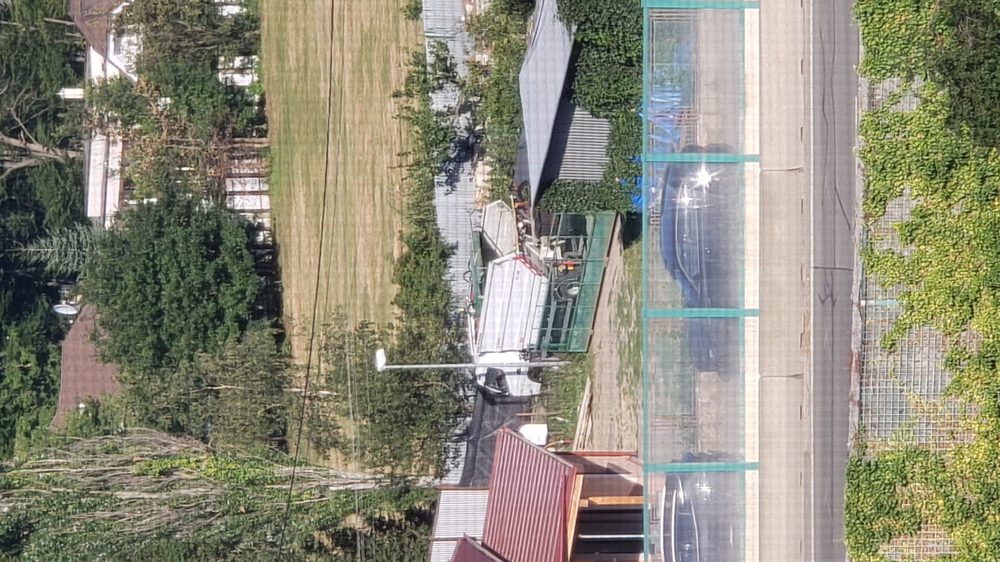 This image depicts a street scene in Prešov, Slovakia. In the foreground, a chain-link fence covered in green ivy separates the viewer from a road. Beyond the fence, two black cars are visible, parked behind a transparent barrier. Further back, a construction site is apparent, with a white truck equipped with a large mechanical arm and a silver-colored trailer. Adjacent to the truck is a small building with a red-tiled roof. The background features residential buildings, trees, and a grassy area. The overall impression is one of a suburban or semi-industrial area on a sunny day.
