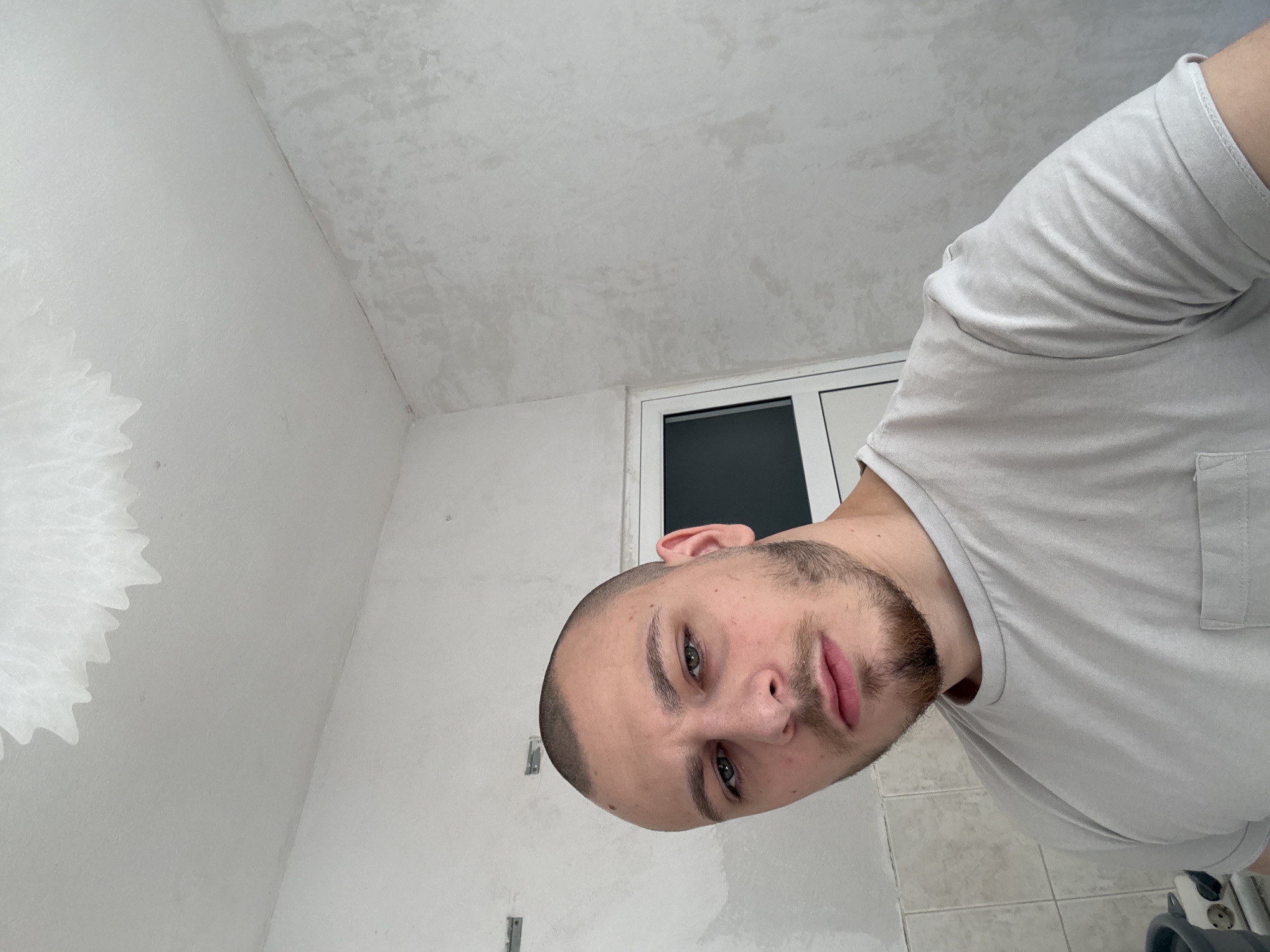 A close-up, tilted selfie shows a man with short, dark hair and a beard in an interior space. The walls and ceiling appear to be white and possibly newly plastered or painted. A window frame is visible in the background, with what appears to be dark glass or covered pane. Based on other context, it is implied the location is Stara Zagora, Bulgaria.