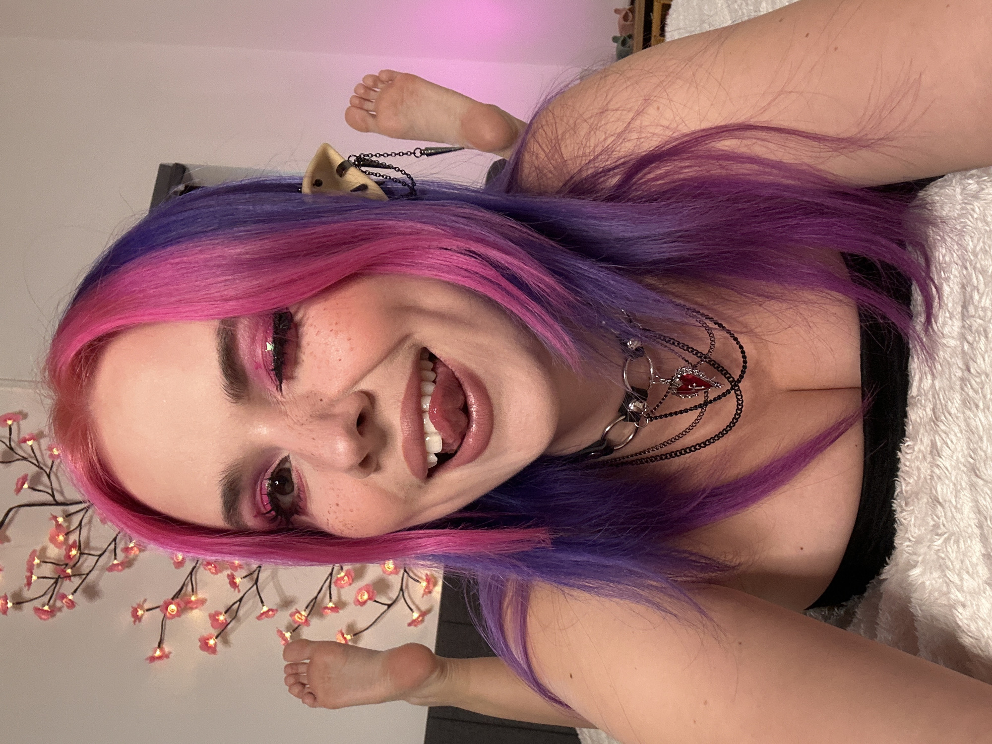 A young woman with pink and purple hair is lying on her back on a white textured surface, possibly a bed or blanket. She is smiling and looking at the camera. She is wearing a black top and a layered necklace with a red jewel. She also has a small object attached to her hair, possibly a charm or decoration. In the background, there are decorative branches with small lights and a pinkish glow. The location cannot be determined from the image.