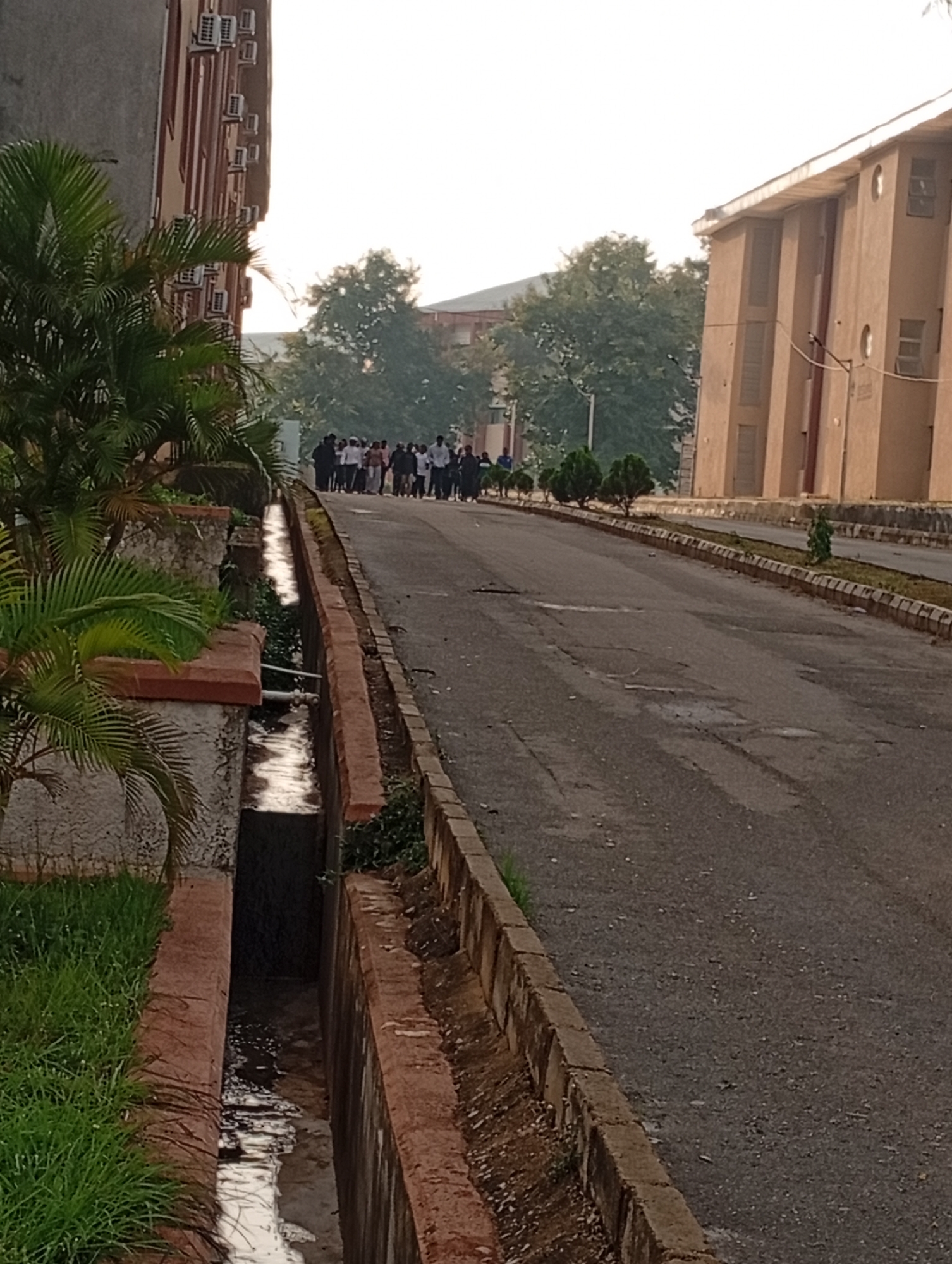 The image captures a view of a paved road leading away from the viewer, flanked by buildings and vegetation. On the left, a building with multiple air conditioning units is visible, partially obscured by lush green foliage. Adjacent to this building is a drainage channel filled with water, bordered by red bricks. On the right, a tan-colored building with several windows and doors stretches into the distance. A group of approximately fifteen people, dressed in casual attire, are walking away from the camera along the road. The road surface is asphalt and shows signs of wear and tear, with cracks and patches. The environment suggests an outdoor setting, possibly a university campus or institutional grounds, given the architecture and the orderly arrangement of the greenery. The lighting indicates it might be morning or late afternoon. This scene is located within the city of Jos, Nigeria.