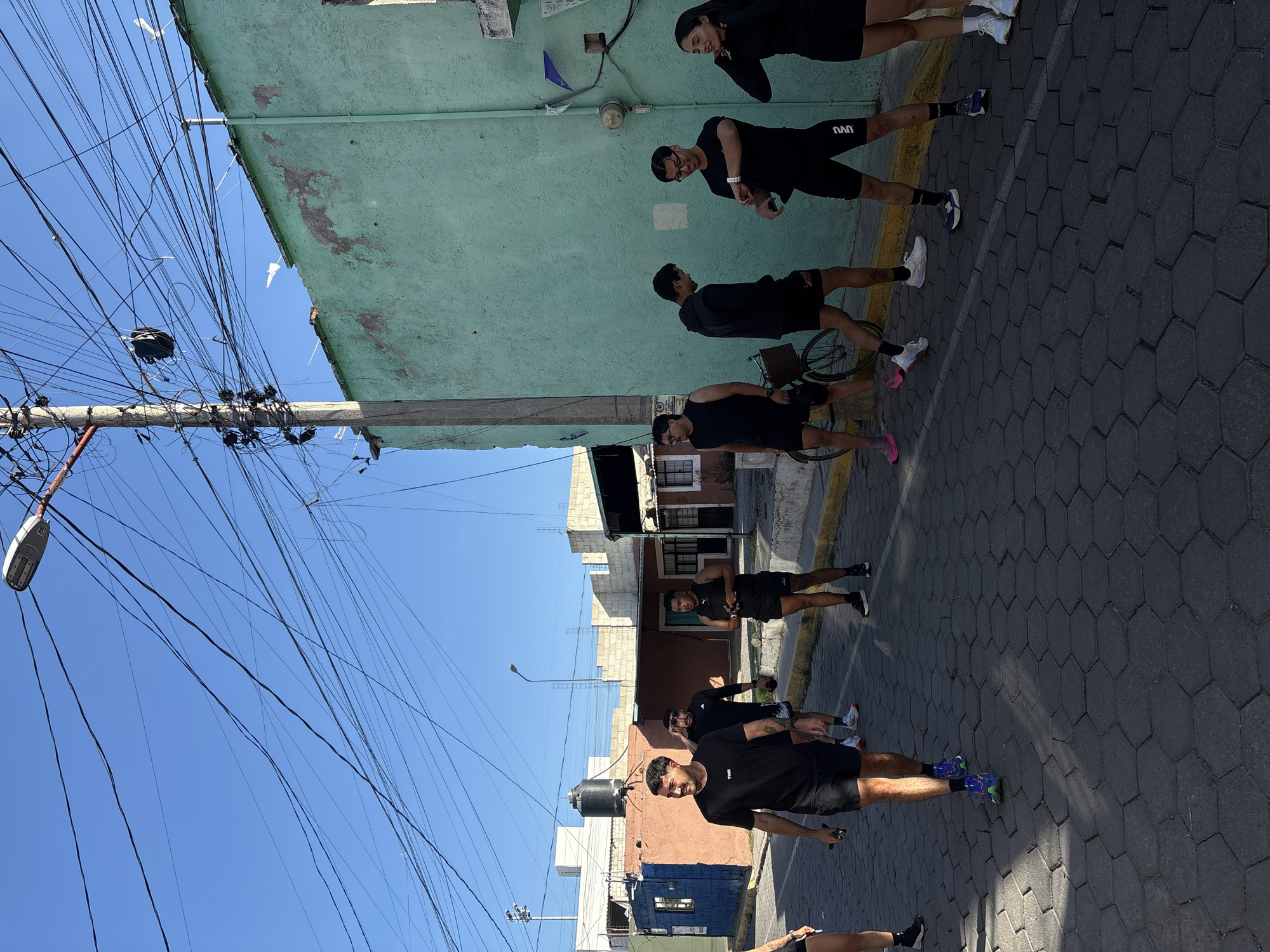 The image shows a group of eight individuals in black athletic wear standing on a cobblestone street in San Andrés Cholula, Mexico. They are aligned roughly in a row along the street, with the first person closest to the camera facing forward and smiling. The others are oriented more towards the side. Behind them is a colorful assortment of buildings with a predominantly light green structure, and a blue sky is visible above, crisscrossed with many electrical wires and a street lamp on the left. A bicycle is next to one of the individuals.