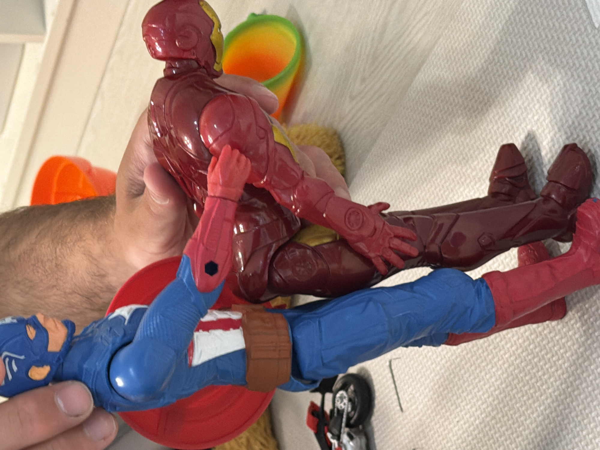 A close-up, vertically oriented photograph captures a person's hands holding two superhero action figures on a textured white surface, possibly a towel or blanket. The scene is set indoors, in Sofiivska Borshchahivka, Ukraine.
The primary subjects are action figures of Iron Man and Captain America. Iron Man, dressed in his iconic red and gold armor, is held by a right hand above Captain America, who lies face-up on the white surface. Captain America is in his blue, red, and white costume, with his red circular shield visible beneath him. The person's left hand is also visible near Captain America's midsection. The pose of the figures suggests a dynamic interaction or play scenario, perhaps a confrontation between the two characters.
In the background, an orange plastic funnel-like object and another toy with green and yellow elements are partially visible, suggesting a play area. A small, dark toy motorcycle or vehicle is also discernible near Captain America's feet. The hands holding the figures have visible hair on the forearm, indicating an adult or older child. The lighting appears to be ambient indoor light, and no specific time of day or weather is evident. There is no visible text in the image.