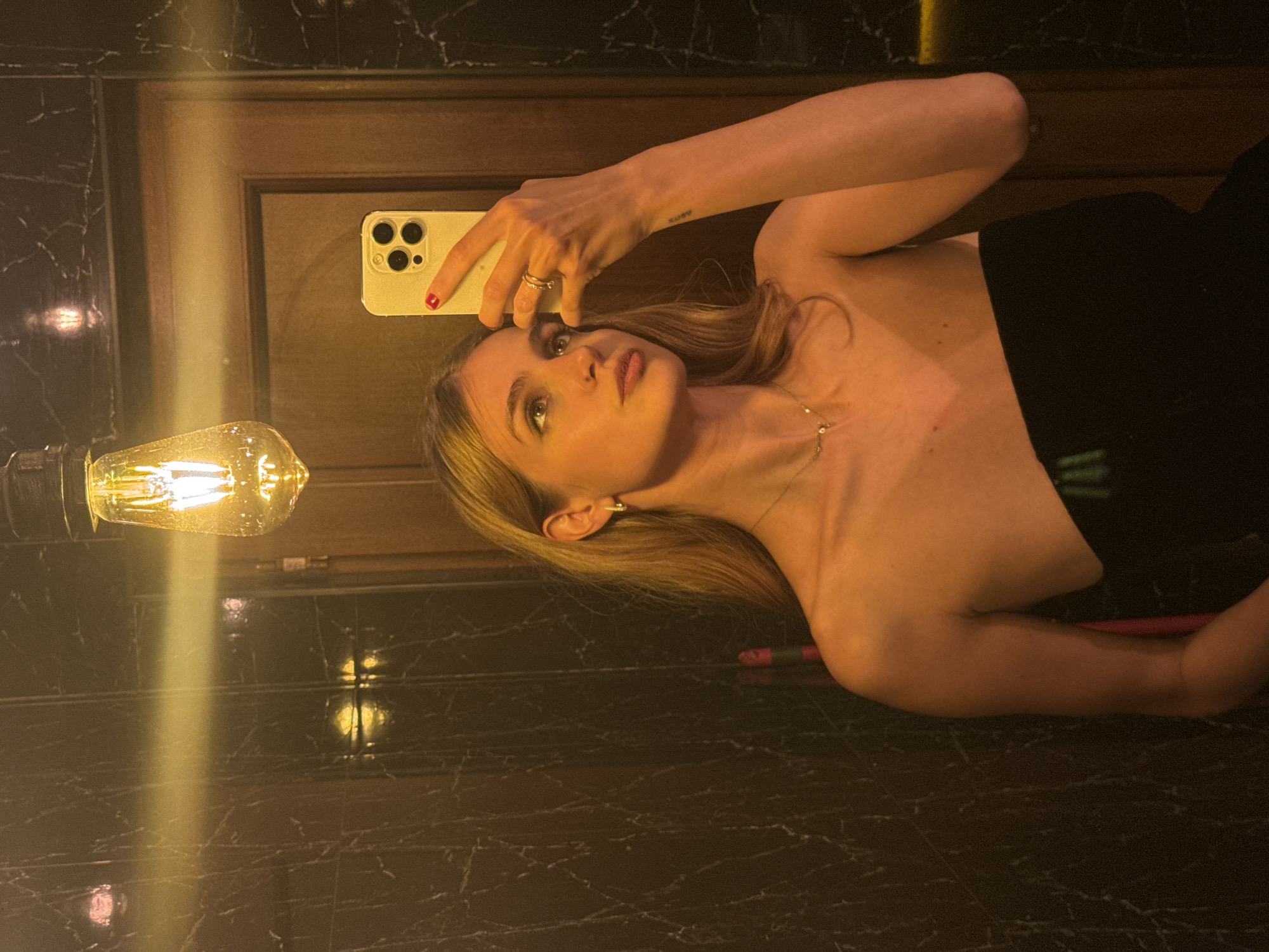 A woman takes a selfie in a mirror. She has blonde hair and is wearing a strapless black top and a necklace. She is holding a cell phone in her left hand. The mirror is in a room with marble walls and a wooden door. An Edison bulb hangs from the ceiling. The room is dark, and the only light source is the bulb. The location is likely a room in Yerevan, Armenia.