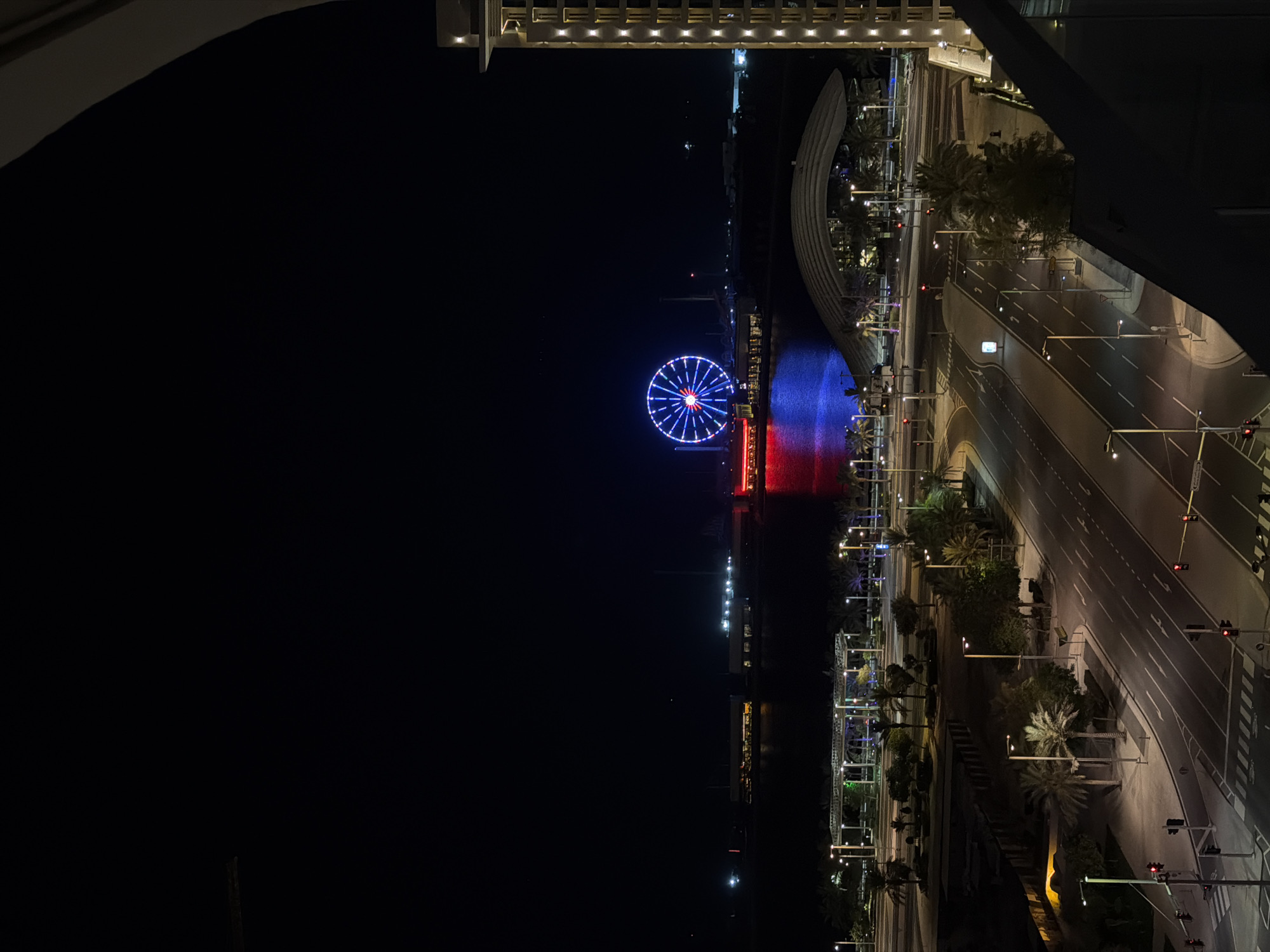 An aerial, nighttime, long shot captures a brightly lit Ferris wheel against a dark sky in the distance. The Ferris wheel, with red and blue lights, is positioned over a body of water. Buildings with illuminated windows are visible along the waterfront. In the foreground, a multi-lane road with white lane markings curves through a treed area with palm trees. Streetlights illuminate the road and sidewalks. A modern building with vertical illuminated windows is on the right side of the image, and a curved architectural structure is visible in the middle ground. Traffic signals show red lights at an intersection. A sliver of an angled dark structure occupies the bottom right corner, suggesting the viewpoint is from within a building.