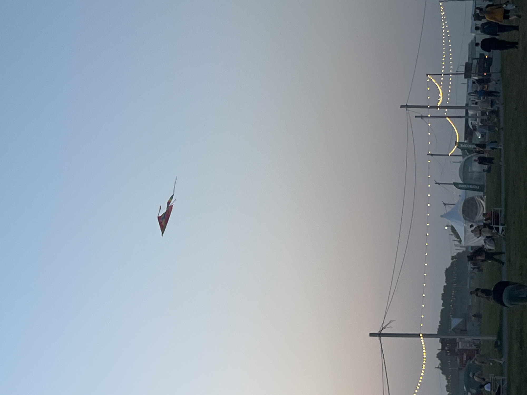 A red kite is flying high in a pale blue sky. Below, a grassy field is dotted with people, tents, and structures. String lights are strung between poles, casting a warm glow across the scene. The overall atmosphere suggests an outdoor event or festival. The location is the city of Surakovo, Russia.