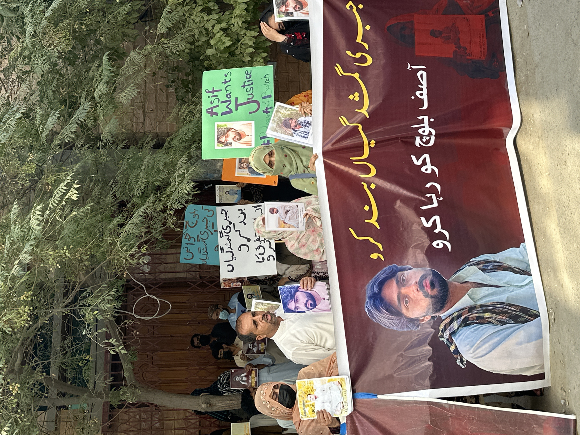 A group of people are holding signs and a banner during a protest in Quetta City Tehsil, Pakistan. The banner, which is red with yellow and white text, features a large image of a man with a beard and a light blue shirt. The text on the banner appears to be in Urdu and translates to "Stop forced disappearances" and "Release Asif Baloch". Some individuals are holding up smaller signs with pictures of men and text in Urdu and English. One sign in the background reads "Asif Wants Justice". The individuals are standing in front of a building with a brown metal gate and some foliage. The ground is a mix of dirt and gravel. The protest appears to be peaceful.