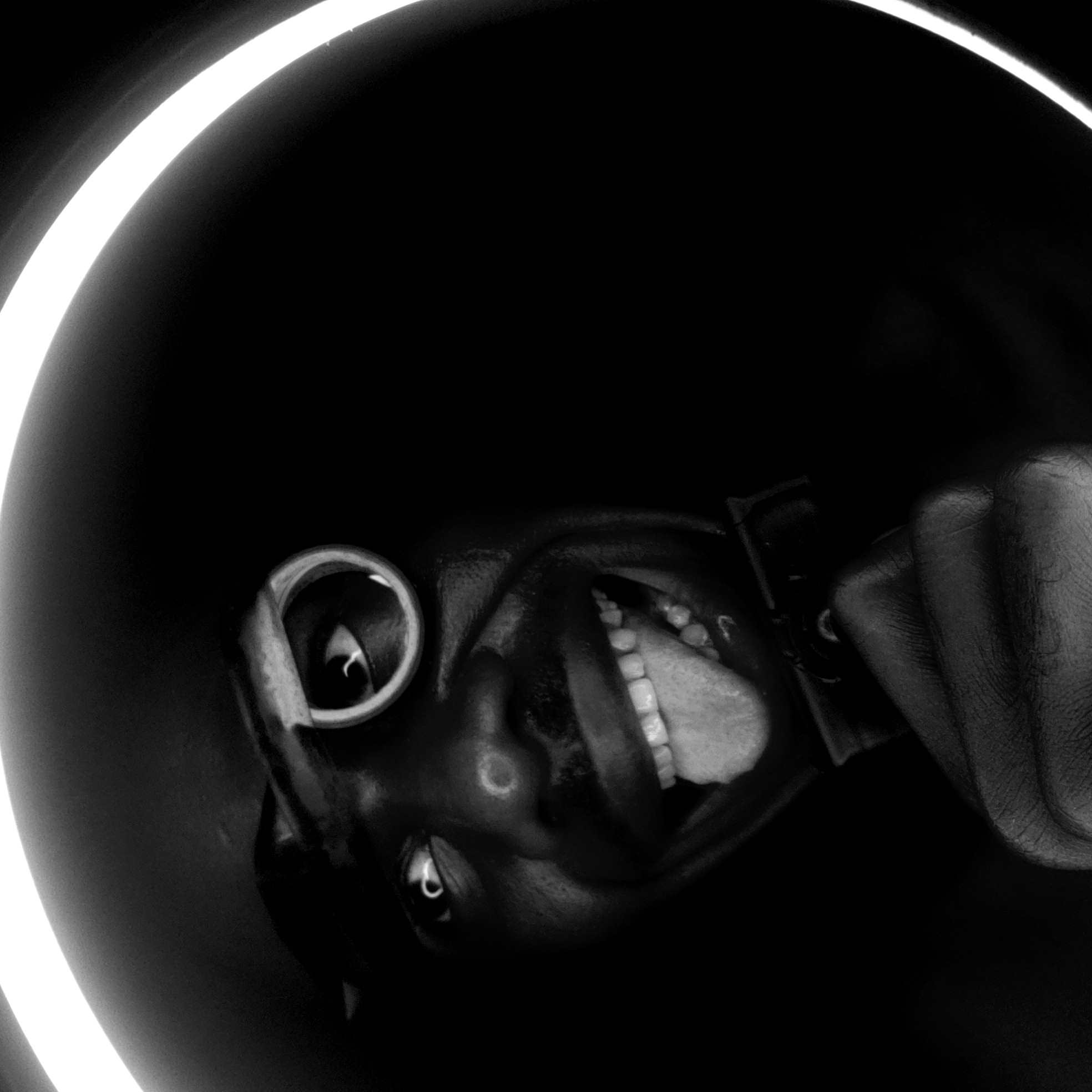 A close-up, black and white photo taken indoors shows a person, likely in Narayi, Nigeria, from an extremely low angle with the camera pointed upward. The person's face is contorted in a wide, open-mouthed expression with the tongue visible. The person is wearing a ring on their left ear. The hand of the person is holding what appears to be a camera or recording device, The face and hand are framed by a circular light source, possibly a ring light, creating a high-contrast image. The background is dark and completely devoid of any other objects or features.