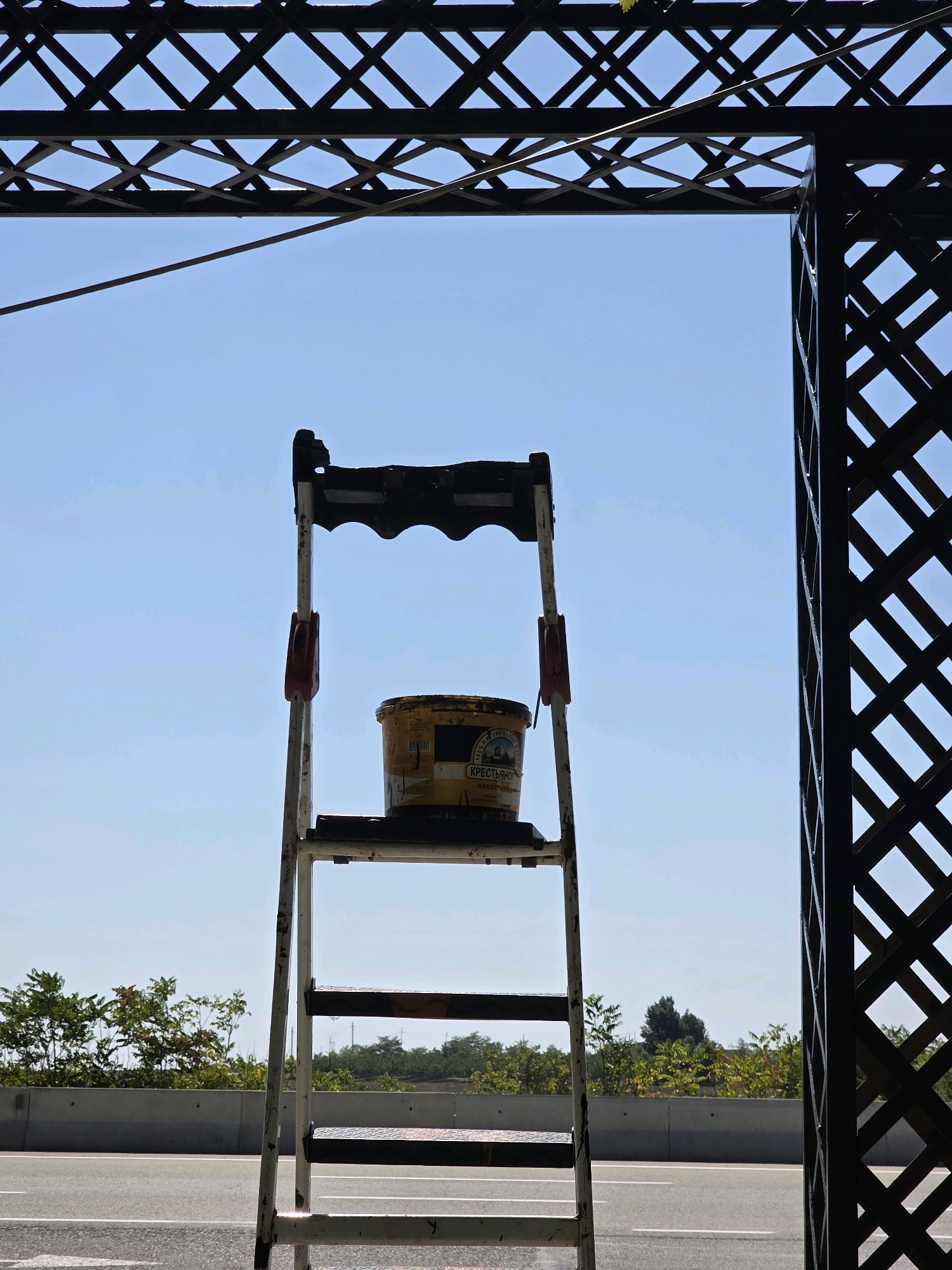 The image, taken in Qobustan, Azerbaijan, depicts a portable stepladder positioned in front of a clear blue sky, framed by dark, lattice-like structures. A yellow bucket with Cyrillic labeling is placed on one of the ladder's steps. In the background, a highway is visible with a concrete barrier, and beyond that, a line of trees and vegetation. The overall impression is of an outdoor scene, possibly near a road construction or maintenance area, with the sky and framing structures suggesting an overhead element like a bridge or canopy.