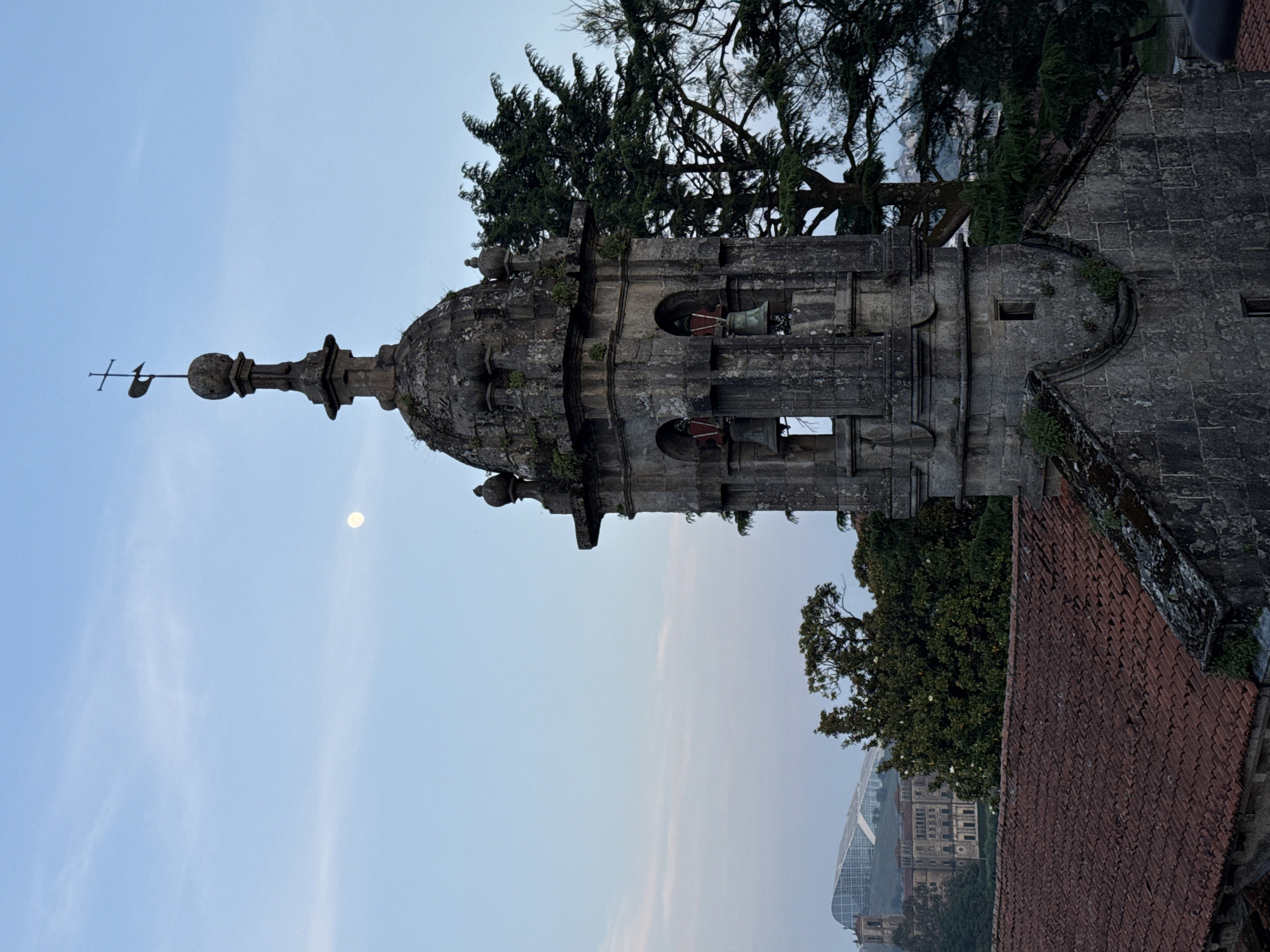 A vertical, full-frame photograph shows a stone bell tower, covered with some plant life, against a light blue sky, taken in Santiago de Compostela, Spain. The top of the tower is round, then the tower continues in a squared design. Small bells are visible in window-like openings. A metal object sits on the very top of the tower. A tree is behind the tower. To the right is a tiled roof. In the distance is a glass building.