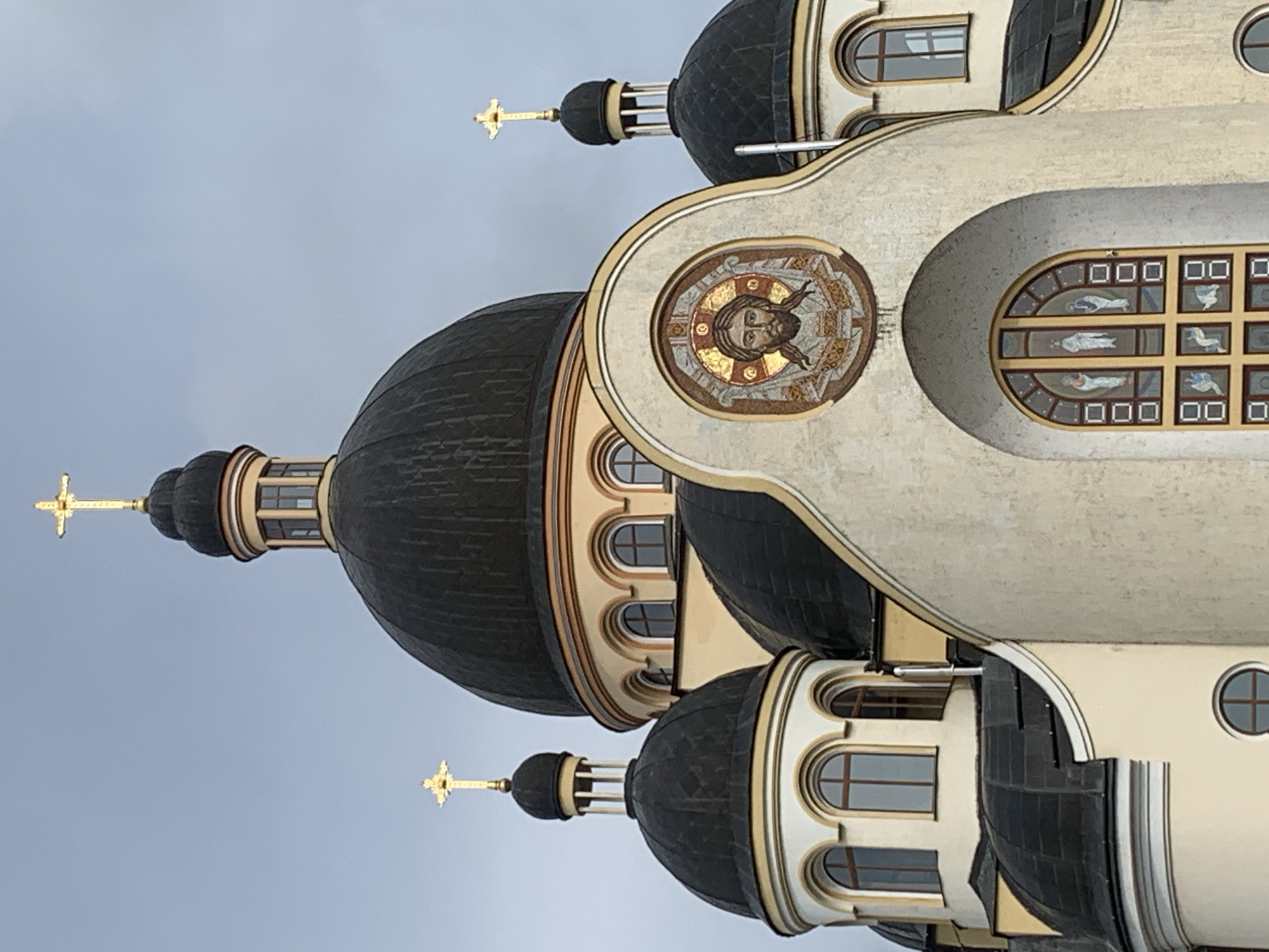 The image shows the exterior of a domed church with a mosaic of Jesus Christ above a large arched stained-glass window. Three golden crosses top three of the church's domes. The church appears to be made of light-colored stucco and has dark domes. The sky is overcast. The church is the Church of the Holy Ascension of Christ in Kolomyia, Ukraine.