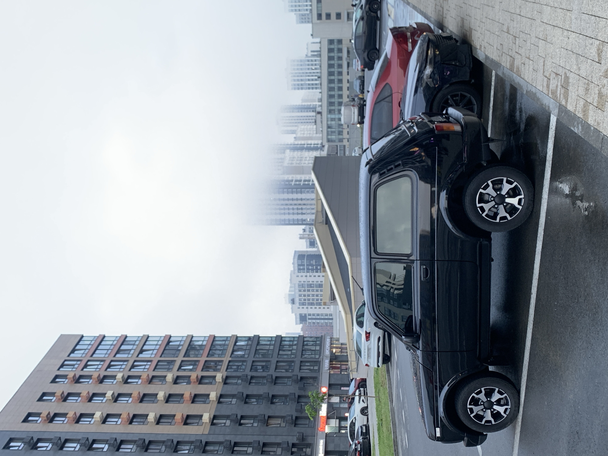In Stavropol, Russia, a black Lada Niva automobile is parked in a wet, paved parking lot. The car is positioned in the foreground, facing left, and its front section is visible. Behind it, a red sedan is parked, followed by a black car with a modified front bumper. Further back, several other vehicles and a white minivan are observable. In the background, a large, modern apartment building stands tall, with numerous windows and balconies. Beyond the apartment building, more residential buildings and a high-rise structure are visible, partially obscured by fog. The sky is overcast, contributing to the gloomy atmosphere of the scene.