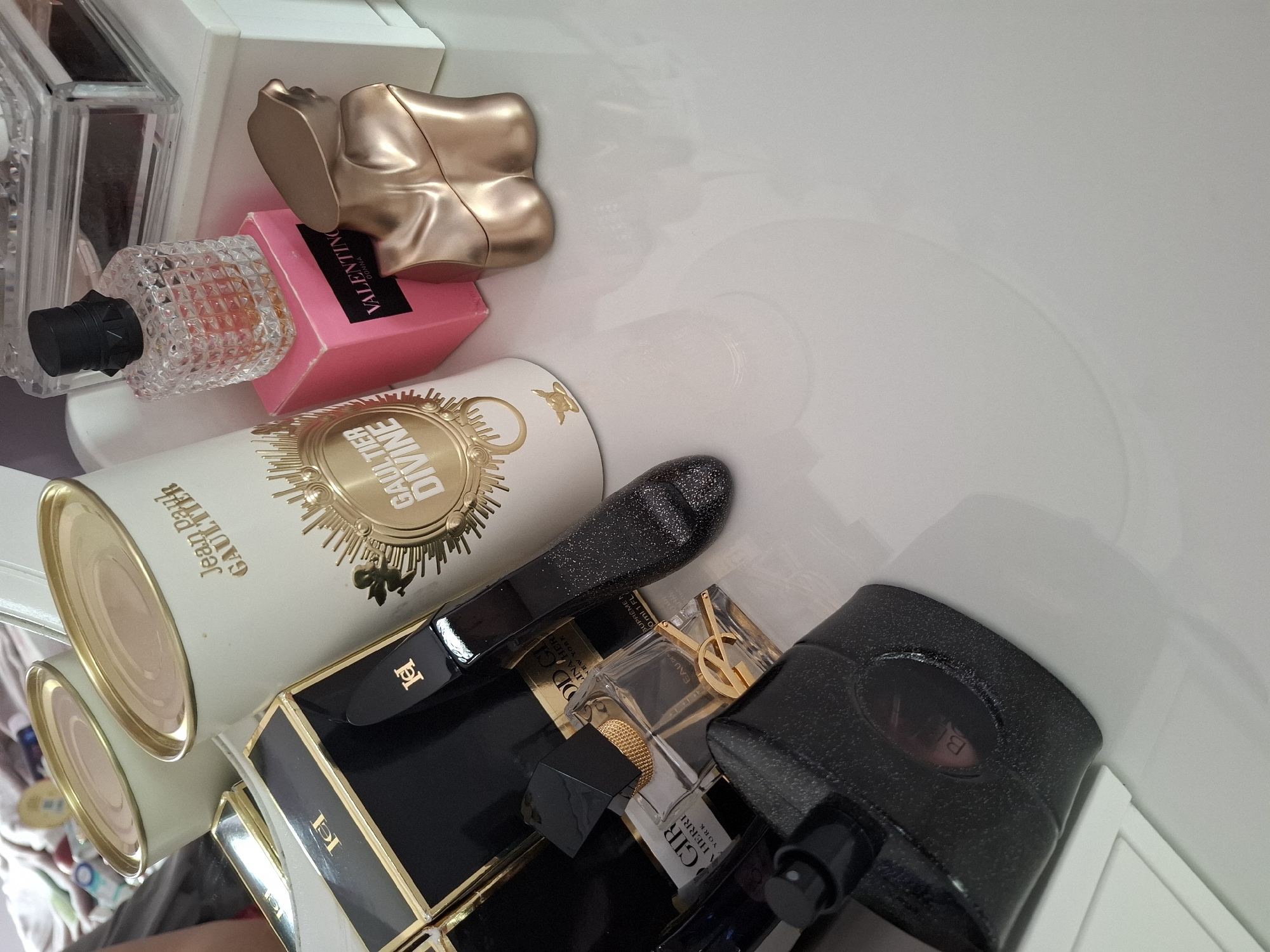 The image shows a collection of perfume bottles and packaging. In the upper left corner, there is a clear glass bottle with a black cap and a pink box labeled "VALENTINO DONNA." Next to it is a gold-colored torso-shaped bottle. A white can with gold trim, labeled "Jean Paul GAULTIER DIVINE," is positioned below. Towards the bottom right are several dark-colored bottles, one with a gold "YSL" logo and another labeled "A HEARRE NEW YORK." The scene appears to be within a white container or drawer. The location, Panagyurishte, Bulgaria, is irrelevant as it does not feature in the image.