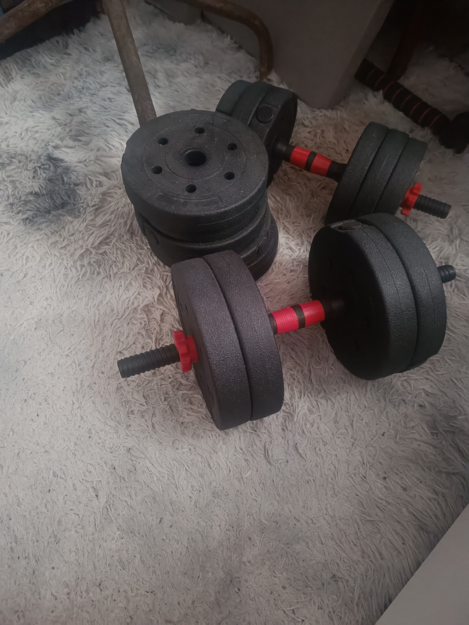 The image shows a collection of adjustable dumbbells lying on a plush, light-colored rug. The dumbbells are black with red collars securing the weight plates. One dumbbell is assembled and appears to be in use, with two weight plates on each side of the handle. Another dumbbell is partially assembled, with its handle and collars visible. A stack of loose weight plates is also present in the background. The scene is indoors, with a wooden object, possibly furniture, visible in the upper left corner. There is no clear indication of the time of day or weather. The lighting is dim, suggesting an indoor setting. The image is focused on the dumbbells and does not contain any people or discernible text. The overall impression is one of a home gym or fitness equipment storage area.