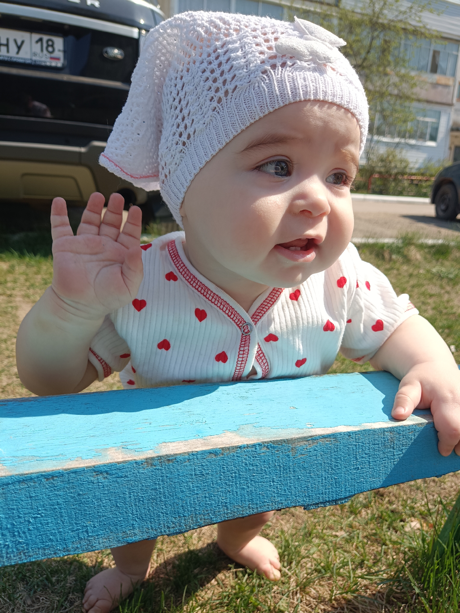 A baby stands next to a blue bench on a sunny day. The baby wears a white, crocheted hat and a white shirt with small, red hearts. The baby's left hand is raised in a waving gesture. The baby is barefoot on a grassy lawn. A black vehicle with a license plate that reads "НУ 18" is parked behind the baby. In the background, a multi-story building is visible. This image was taken in Noviy, Russia.