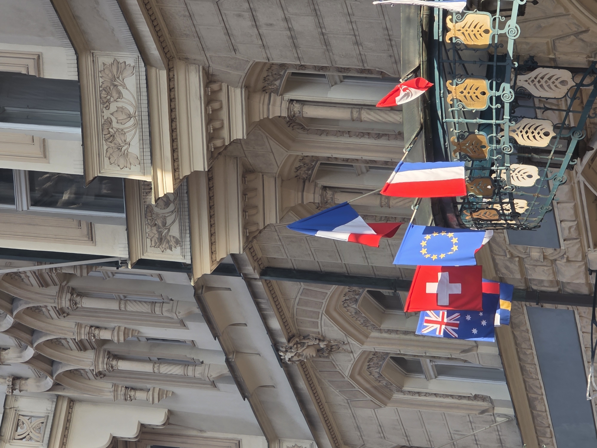 The image captures a vibrant street scene dominated by ornate European architecture and a display of multiple national flags. The perspective is looking upwards towards the facades of several multi-story buildings, which are richly decorated with classical elements.
The buildings feature intricate cornices, decorative moldings, and carved details. Elaborate floral patterns are visible on friezes, and some sections showcase arched structures and fluted or spiraled columns. The architecture suggests a historical and grand style, likely found in an old European city. The windows are tall and framed, reflecting parts of the surroundings.
A decorative metal balcony railing with a distinctive fish-shaped motif runs across one of the buildings, adding a unique character to the scene. From this balcony, or possibly from poles attached to the building, several flags are prominently displayed, fluttering in what appears to be a gentle breeze.
Visible flags include the blue, white, and red tricolor of France, the blue flag with twelve golden stars of the European Union, the red flag with a white cross of Switzerland, and the blue flag featuring the Union Jack and the Southern Cross constellation, representing Australia. A smaller red and white flag is also visible, and another blue and yellow flag is partially obscured.
The lighting indicates a bright, clear day, casting subtle shadows on the architectural details and illuminating the colorful flags. There are no visible people or discernible text in the image. The arrangement of flags suggests a festive or international atmosphere in this elegant urban setting.