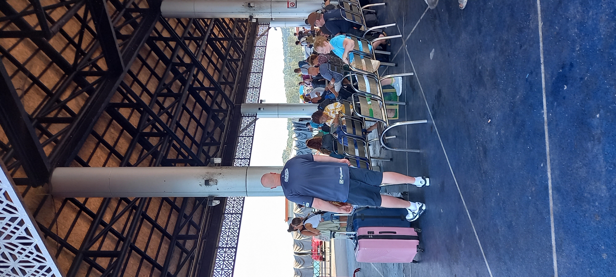 This image captures a scene from a waiting area, likely a ferry terminal, given the context of Thong Sala, Thailand. The photo is oriented sideways but depicts a covered outdoor space bustling with people.
In the foreground, a bald man stands facing away from the camera. He wears a dark blue polo shirt with "SEKO" prominently displayed on the back and "MyGames" visible below, along with dark shorts and white sneakers. Next to him are two pieces of luggage: a bright pink hardshell suitcase and a darker blue one.
Beyond him, several rows of people are seated on simple metal and plastic chairs, facing forward. They appear to be waiting, possibly for boarding. Various individuals are visible in casual attire, including someone in a light blue top and another in a yellow patterned shirt.
The waiting area is covered by a large, intricate roof structure made of dark metal trusses, providing shade. This roof is supported by several large, light grey cylindrical pillars. In the background, beyond the seated passengers, a bright outdoor scene is visible, suggesting clear weather. This area shows glimpses of water and distant green land, consistent with a port setting. A red and white "No Smoking" sign is visible on one of the distant pillars. The ground is a dark blue, possibly painted concrete or tarmac, with white lines marking sections. The scene suggests a busy transit point during daytime.
