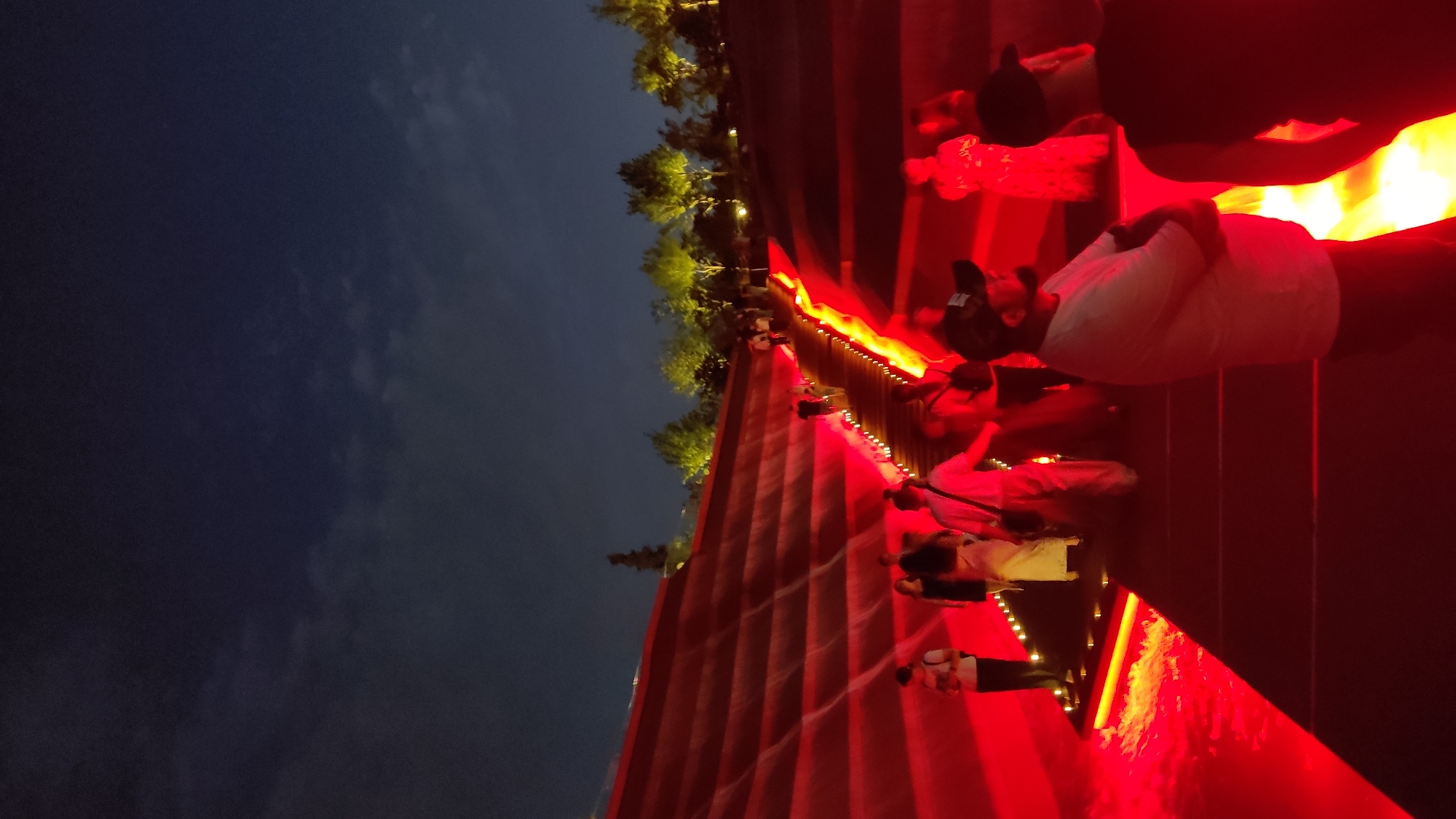 The media file shows a nighttime scene with people gathered on tiered red steps, illuminated by red lights and some yellow-orange lights. The sky above is dark and cloudy, suggesting it is evening or night. Trees with green foliage are visible on the left side of the frame, hinting at an outdoor location.
The people are mostly seen from the back or side, and their activities are not clearly discernible, though they appear to be standing and perhaps observing something. One person in the foreground is wearing a white shirt. The overall atmosphere is festive and vibrant due to the strong red lighting. There is no visible text in the media file. The location context is Krasnodar, Russia, and the scene might be a public space or an event venue utilizing dramatic lighting.