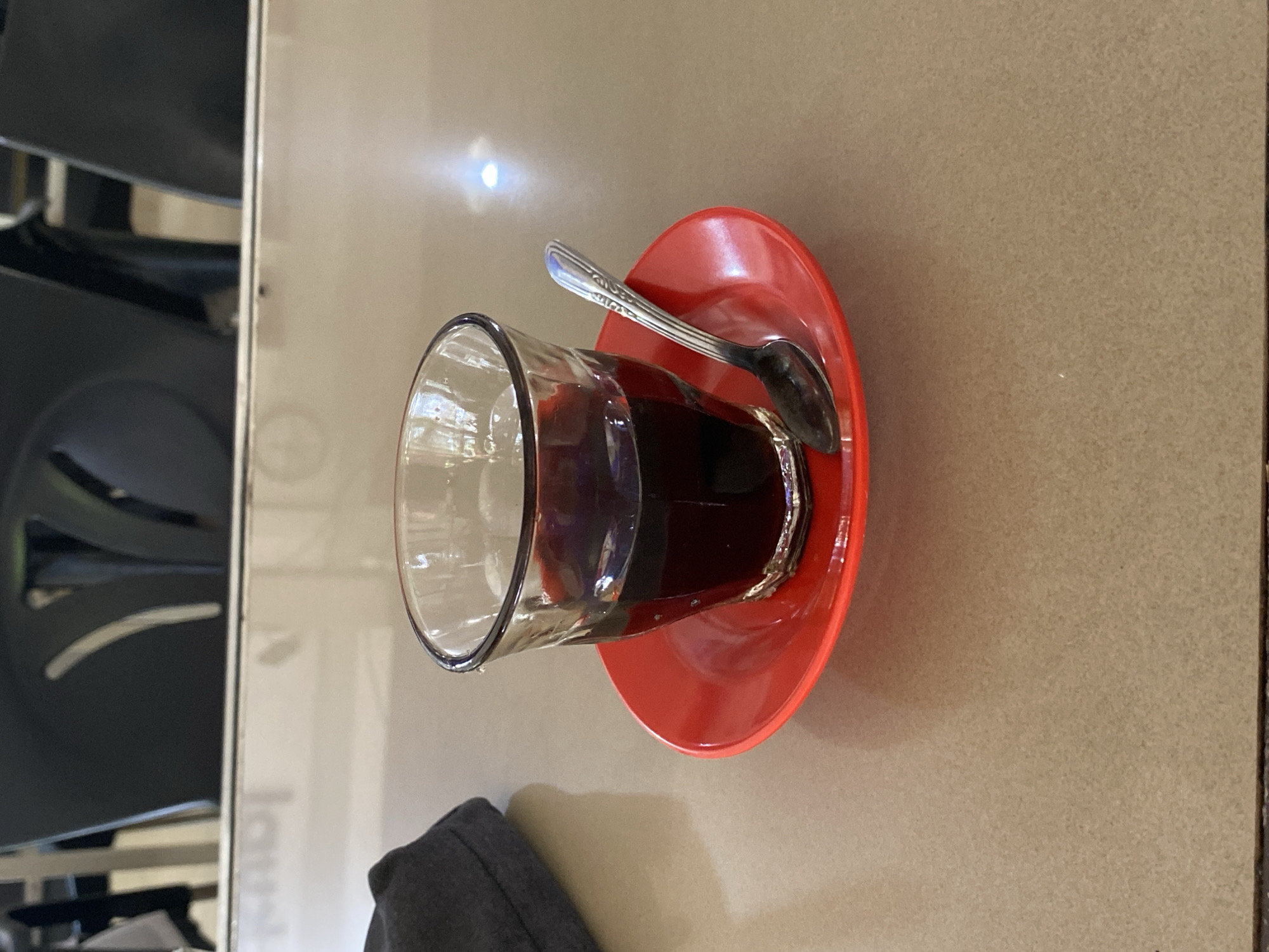 A glass of dark liquid, likely coffee or tea, sits on a red saucer on a light brown table. A silver spoon rests on the saucer. In the background, blurred black chairs are visible. The scene appears to be in a cafe or restaurant. The location is identified as Lhokseumawe, Indonesia.