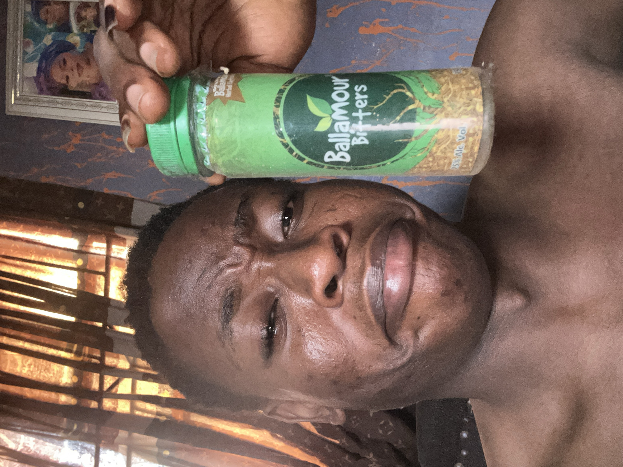 A man is holding a bottle of "Ballamour Bitters" over his head indoors. The bottle is green with a green cap and has the words "Ballamour Bitters" written on it. The man is dark-skinned and has a displeased expression on his face. The background is slightly blurred and appears to be an indoor space. The location is not specified.