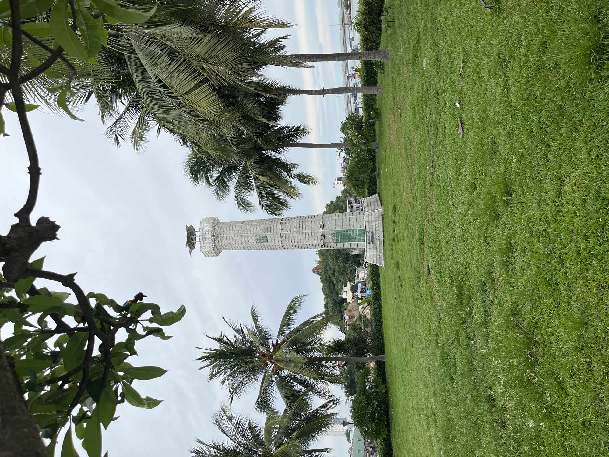 In Nha Trang, Vietnam, a white lighthouse stands tall in a grassy park, framed by lush green palm trees. The lighthouse is a cylindrical structure, wrapped in a white lattice-like material, with a decorative element at its peak. In the background, the city skyline rises behind a hill adorned with a historic tower. Boats are visible on the water, and a bridge spans the bay, hinting at the coastal setting. The sky is overcast, casting a soft, diffused light over the scene. The foreground features a well-maintained lawn with scattered fallen leaves and some debris. The overall impression is one of a serene and picturesque seaside location.