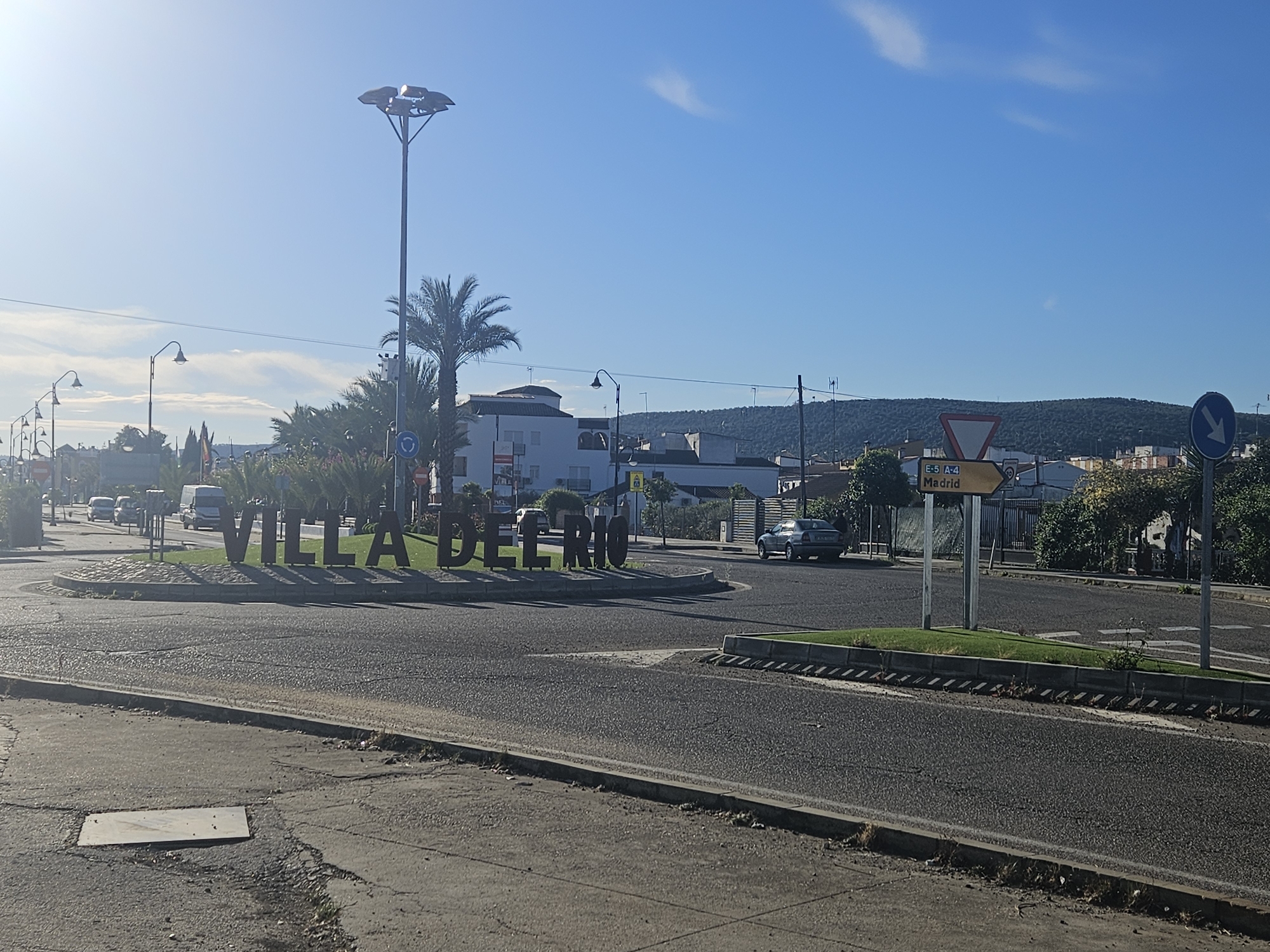 The image shows a street scene in Villa del Río, Spain. A large "VILLA DEL RIO" sign is prominently displayed on a traffic island in a roundabout. Several vehicles are visible on the roads. Streetlights line the road, and a palm tree is situated near the sign. A road sign indicates the direction of Madrid via the E-5 and A-4 highways. Buildings are visible in the background, with a hilly landscape further beyond. The sky is blue with a few clouds. The foreground shows a paved area with a rectangular service box.