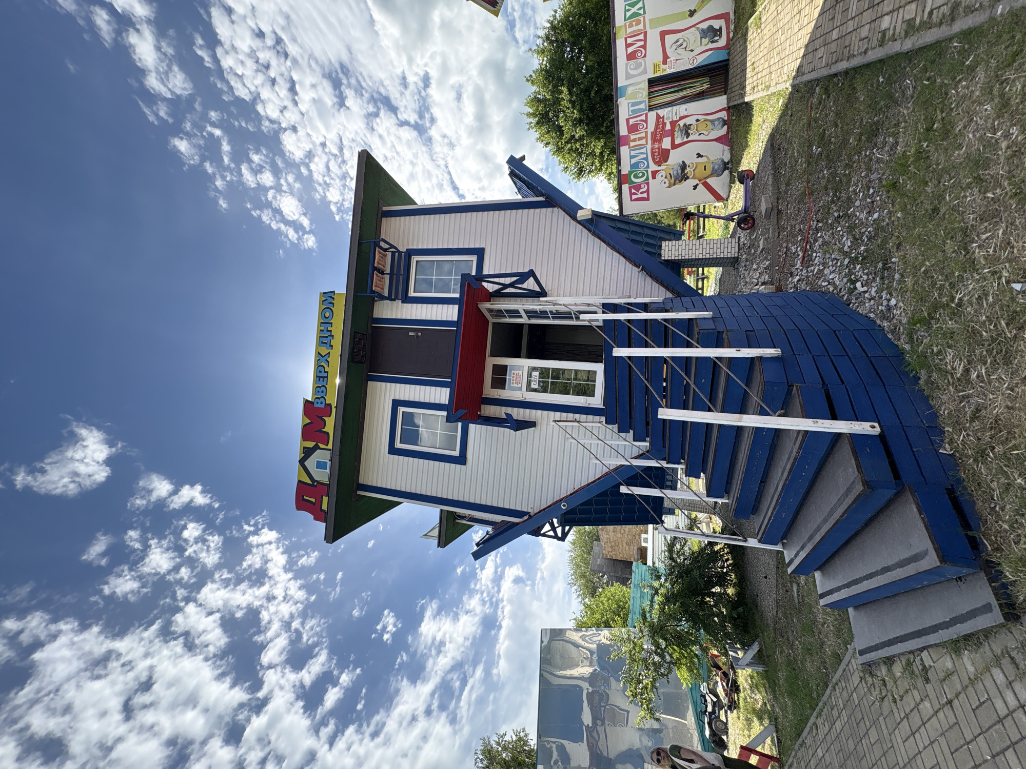 A building called "Inverted House," located in Anapa, Russia, stands on its roof and is tilted slightly to the left. The building is white with blue trim. It has a blue staircase that appears to lead to the second story. A sign in Russian reads "Inverted House." The background contains blue sky, white clouds, a tree, and a sign.