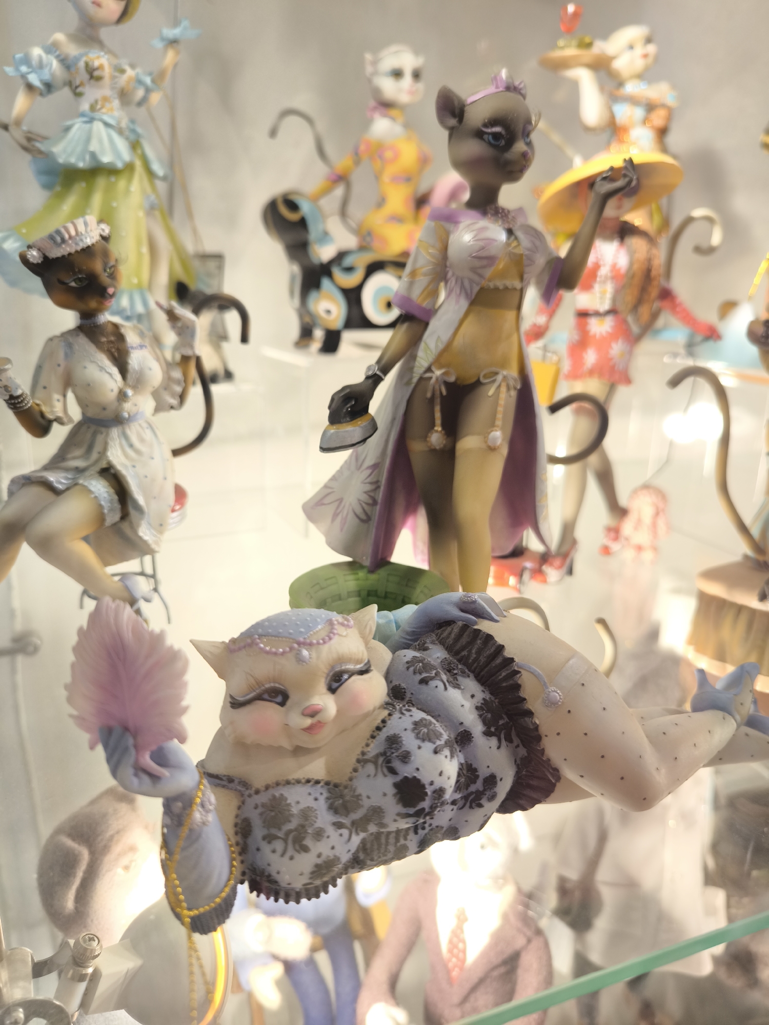 The image depicts an indoor display of multiple anthropomorphic cat figurines, likely situated in Zelenogradsk, Russia, possibly within a cat-themed museum or gallery. The figurines are posed in various human-like activities and attire, presented within a glass display case or on clear glass shelves.
In the prominent foreground, a large, light-skinned cat figurine reclines on its back, head slightly elevated and turned towards the viewer. It wears a patterned light blue bustier with dark trim, light blue gloves, a beaded necklace, and a light blue headpiece adorned with pink pearls. Its left hand holds a pink feather fan. Its right leg is bent, revealing a white stocking with black polka dots, a garter with a purple jewel, and a light blue high-heeled shoe.
To the upper left of the reclining cat, a brown-skinned cat figurine sits on a red-topped stool with a metal base. It is dressed in a white and light blue polka-dotted dress with lace trim and a white headpiece. Its right hand holds a white object. One leg, clad in a white stocking with lace and a white high-heeled shoe, is visible.
Above the reclining cat, a grey-skinned cat figurine stands upright, dressed in a yellow strapless top, dark gartered stockings with white ties, and a white and purple floral-patterned robe. It wears a purple headpiece, and its left hand is raised, holding a black object resembling an iron.
In the mid-ground and background, several other blurred anthropomorphic cat figurines are visible. These include one to the far left wearing a light blue top and a light green skirt, and another to the upper right wearing a yellow wide-brimmed hat and a red floral dress. Additional figures in varied outfits and poses are partially obscured, including one in yellow and orange next to a stylized black and blue dog-like creature, and another holding a tray with a red item. The environment consists of plain, light-colored walls behind the glass display, with reflections visible on the glass surfaces. A metal fixture is present in the lower left foreground. No human individuals are visible in the scene.