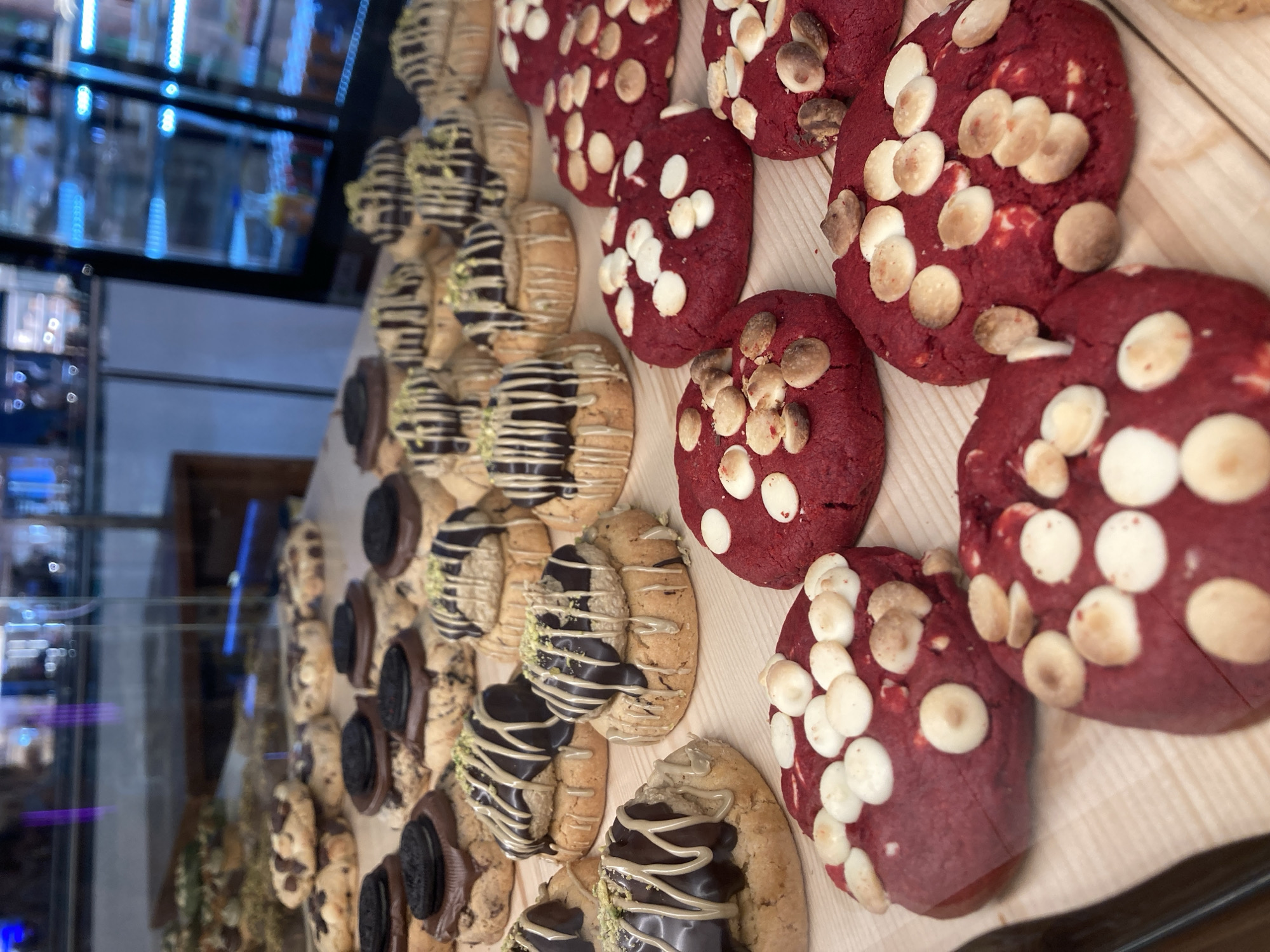 The image displays a close-up, eye-level view of a variety of cookies arranged on a wooden surface, likely in a bakery display case. The cookies vary in flavor and appearance. Some are light-colored with chocolate chips and a dark chocolate drizzle with green sprinkles. Others are red velvet cookies topped with white chocolate chips and macadamia nuts. The background is blurred but suggests a retail environment, with glass cases and reflections of lighting and possibly refrigerators. No people are visible, and no specific location in Barcelona, Spain, is discernible from the image.