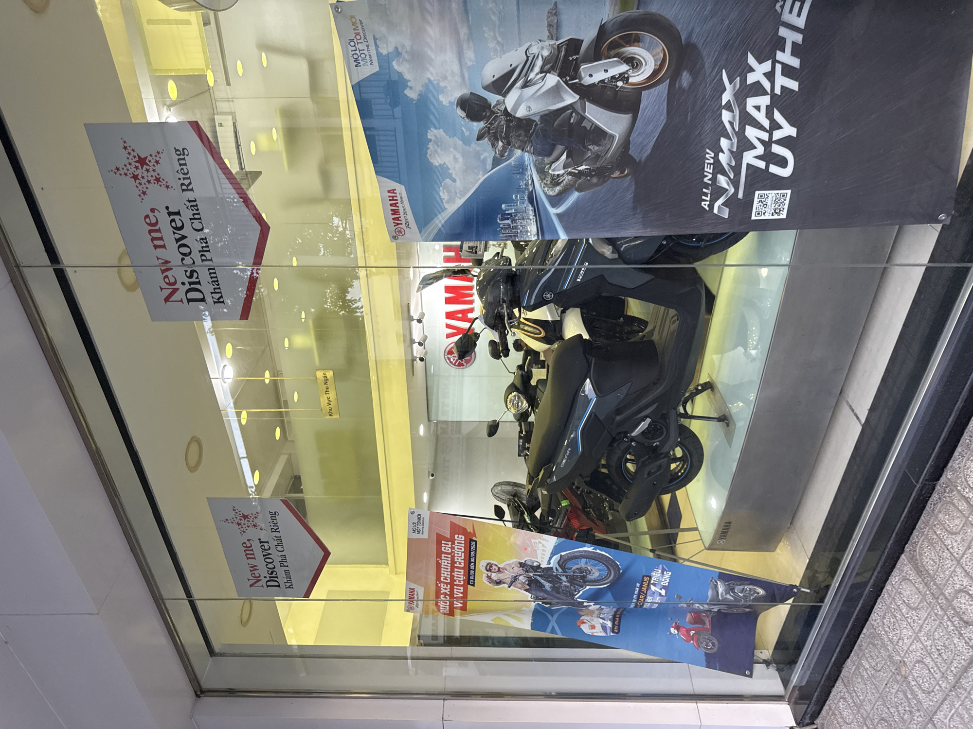 This is a storefront display for Yamaha motorcycles in Nha Trang, Vietnam. The scene features a large glass window with various promotional banners and motorcycles visible inside.
Two triangular banners hang from the ceiling. The banner on the left reads "New me, Discover Khám Phá Chất Riêng" (Discover Your Own Quality) with a red star graphic. The banner on the right also says "New me, Discover" and has a similar red star graphic, with Vietnamese text below.
Inside the showroom, several Yamaha motorcycles are displayed. A prominent banner on the right side of the window features an image of a rider on a scooter with a city skyline in the background. This banner has text including "ALL NEW NMAX" and "MAX UY THỂ". Another banner on the left displays a red scooter and a rider on a motorcycle, with text in Vietnamese.
The interior of the showroom appears to be brightly lit, with yellow lighting visible. Yamaha logos are visible on the motorcycles and a banner inside. A sign that reads "Khu Vực Thu Ngân" (Cashier Area) is visible in the background. The floor inside is tiled, and the exterior sidewalk is made of patterned concrete. The overall impression is of a clean and modern motorcycle dealership.