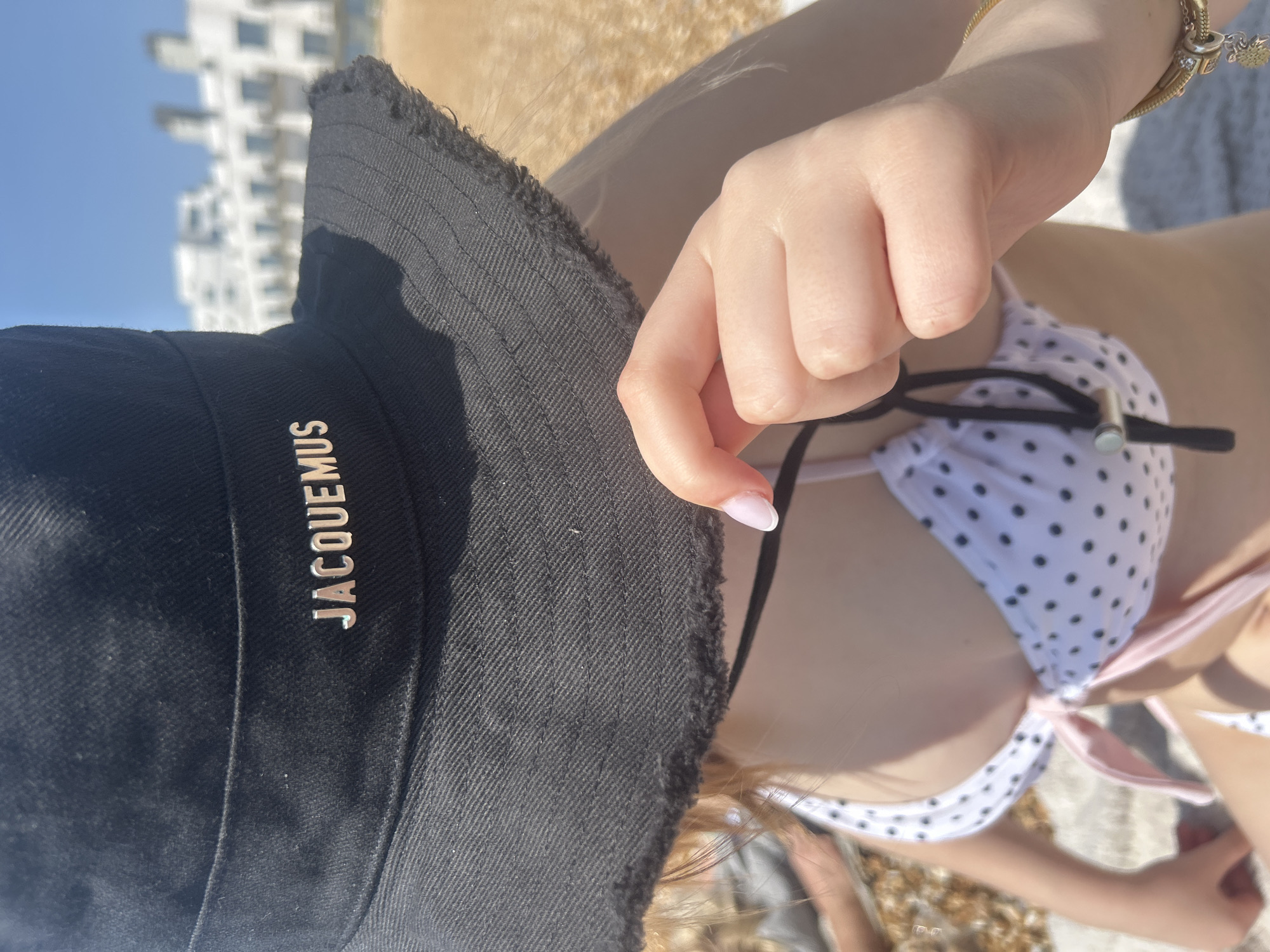 A medium shot of a person wearing a polka dot bikini and a black JACQUEMUS bucket hat lying on a beach in Folkestone and Hythe District, United Kingdom. The person's hand is in the foreground, adjusting the bikini top, and a gold bracelet is visible on their wrist. The background features a blurred view of buildings under a clear blue sky. The beach itself is composed of pebbles and sand. The shot is taken from a high angle.