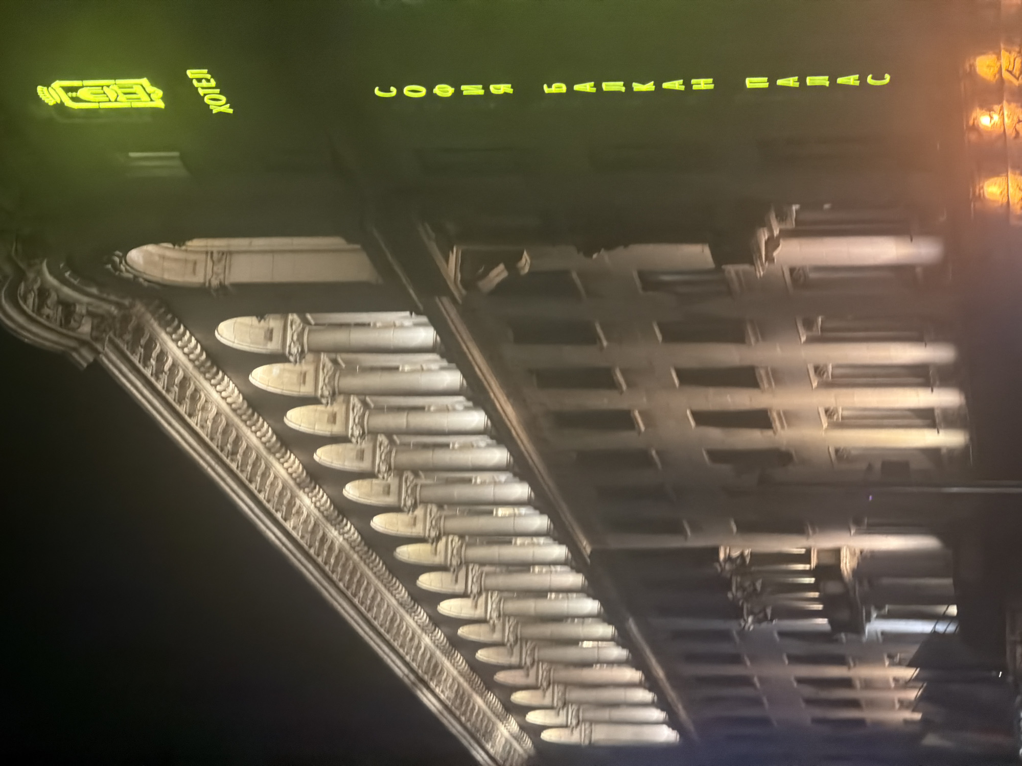 The image captures the illuminated facade of a grand building at night, viewed from a low angle. The architecture suggests a classical or historical style, characterized by a long series of ornate columns or pilasters that line the upper section of the building. These architectural features are prominently lit by upward-pointing lights, creating a dramatic interplay of light and shadow across the detailed stonework.
High on the building's dark facade, neon yellow signs are visible. One sign features a stylized emblem resembling an old lantern or lamp, followed by the Cyrillic word "ХОТЕЛ" (HOTEL). Below this, another line of Cyrillic text spells out "СОФИЯ ПАЛАС" (SOFIA PALACE), clearly identifying the establishment as the Hotel Sofia Palace.
The scene is set at night, with the sky appearing dark. The artificial lighting on the building contributes to a stately and imposing atmosphere. No people or specific activities are visible in the frame, focusing solely on the architecture and its nighttime illumination.