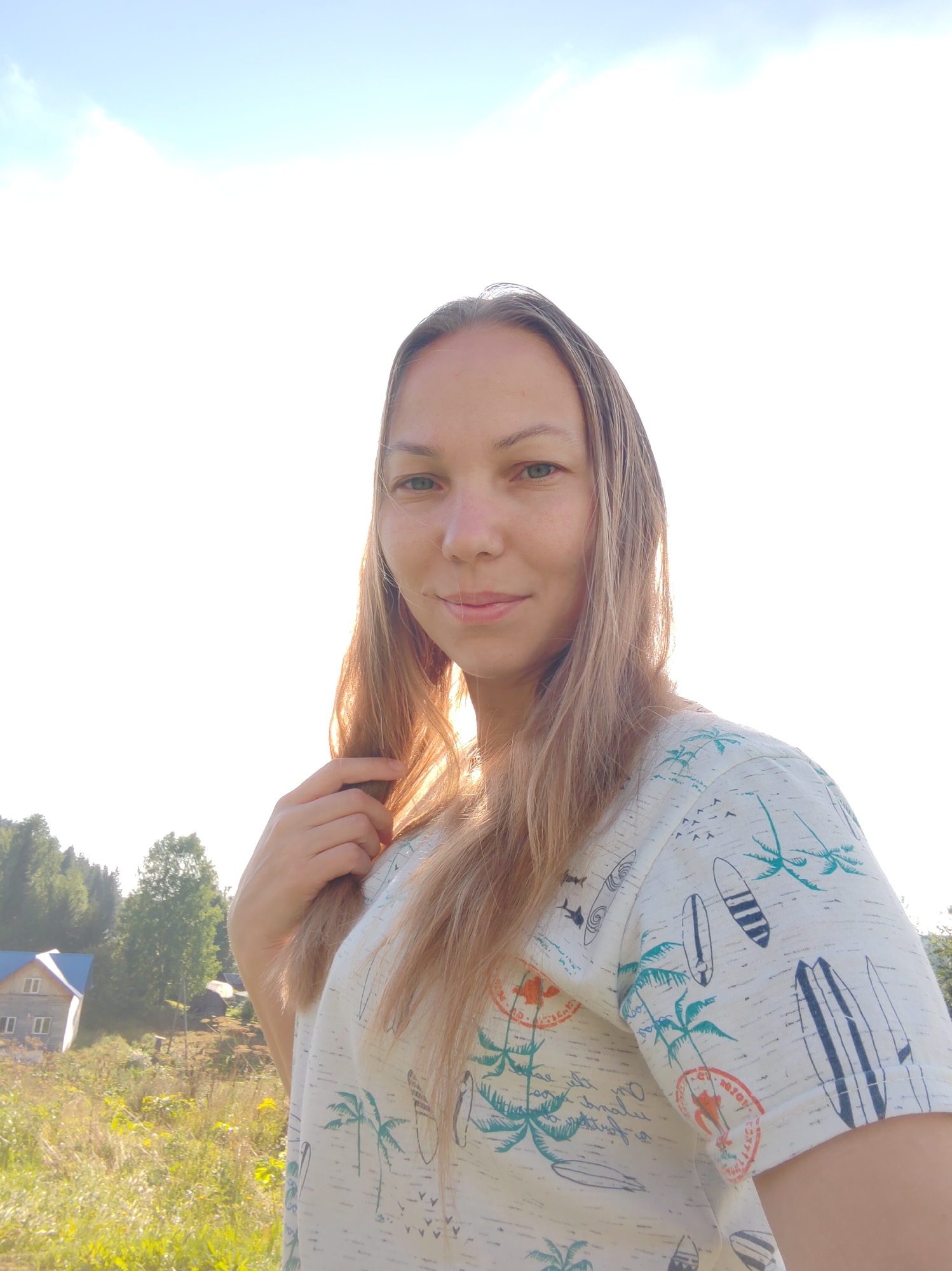 A young woman with long blonde hair, wearing a light-colored t-shirt with a palm tree and surfboard print, is pictured outdoors during the daytime. She is looking directly at the camera with a slight smile. Her right hand is touching her hair. The background shows a sunny sky with some clouds, a grassy field with wildflowers, trees, and a small building with a blue roof on the left side. The overall setting appears to be rural, possibly in or near the city of Chukhlem, Russia, given the architectural style of the building and the natural landscape. The lighting suggests it's either morning or late afternoon.