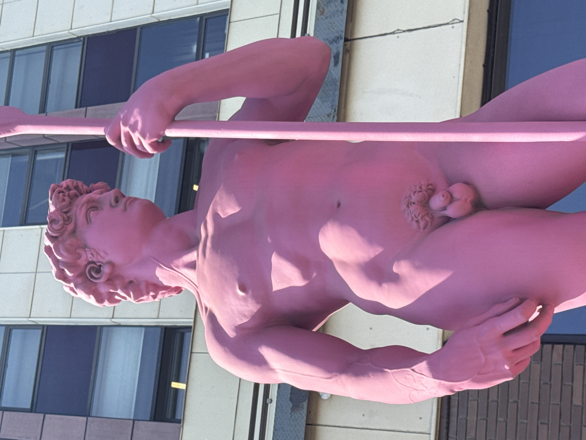 This media file features a large, pink sculpture of a male figure. The sculpture depicts a muscular man holding a pole horizontally across his body. The figure is rendered in a classical style, with detailed musculature and facial features. The sculpture appears to be made of a solid material, possibly stone or metal, and is painted a uniform pink color.
The sculpture is positioned outdoors, with a modern building featuring large windows in the background. The building's facade is light-colored, and the windows are tinted dark blue and reflect the sky. The lighting suggests it is daytime, with strong shadows cast by the sculpture, indicating bright sunlight. The overall scene conveys a sense of public art installation in an urban or semi-urban environment. There is no visible text in the image. The file does not contain any information about the specific activity or event taking place, nor does it provide any explicit emotional cues beyond the static nature of the sculpture. The location context of Zhabkino, Russia, is provided but not visually evident within the frame itself.
