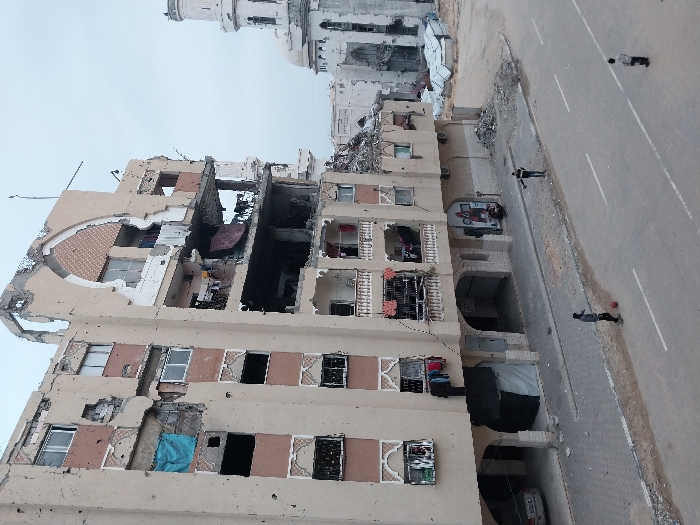 A severely damaged residential building shows gaping holes and structural damage, likely caused by bombing or heavy shelling. Personal belongings are visible within the destroyed apartments. A mosque or significant building is visible in the background. A road runs alongside the building, with several people walking on it. Debris litters the area. The overall scene depicts a war-torn or disaster-stricken urban environment.