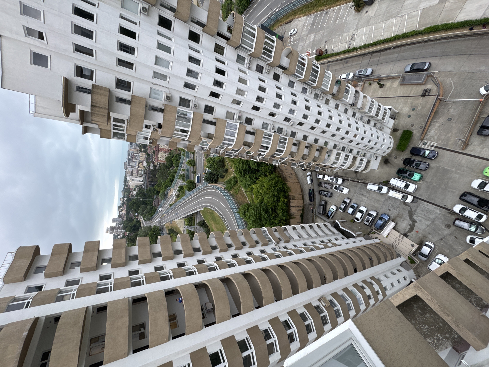 An aerial, slightly angled shot showcases a residential complex in Sochi, Russia, with two tall, white apartment buildings dominating the foreground. The buildings feature numerous balconies with tan railings and a multitude of windows. A multi-lane road curves through the scene in the mid-ground, flanked by lush green trees and other city buildings in the distance. The sky is overcast with gray clouds.
In the immediate vicinity of the buildings, a paved parking lot is visible, with several cars parked in designated spots. Among the vehicles are white, black, silver, and dark blue sedans, as well as some SUVs.
On one of the balconies of the left-hand building, a single person is standing, looking out. No other people are clearly visible or actively engaged in any interactions within the frame. The overall impression is of a densely populated urban environment with a focus on residential architecture and transportation infrastructure.