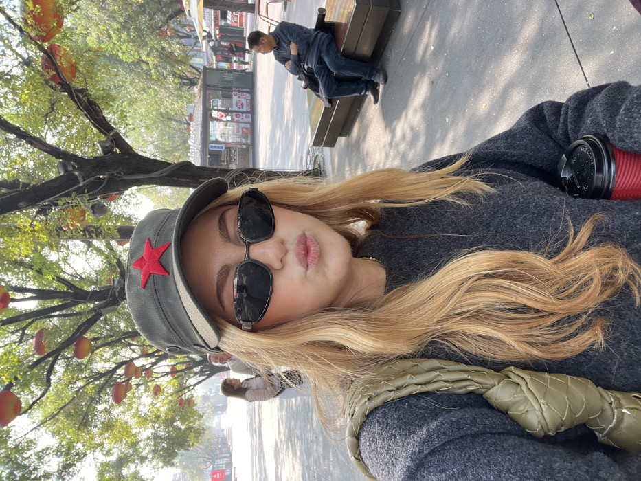 The image depicts an outdoor pedestrian street scene in Yanta District, China. In the foreground, a woman with long, wavy blonde hair is captured in a selfie-style shot. She wears a dark grey, long-sleeved sweater, oval-framed dark sunglasses, and an olive-green military-style cap adorned with a prominent red five-pointed star emblem centered on the front. A braided olive-green shoulder bag is visible on her left side, and she holds a red disposable coffee cup with a black lid in her right hand. Her lips are pursed.
The background shows a paved thoroughfare lined with deciduous trees. Numerous orange-red lanterns are strung between the trees overhead. To the woman's left, two individuals are visible walking away in the distance. To her right, a man in a dark blue long-sleeved shirt and blue jeans is seated on a dark, modern-style bench, with his arms crossed. Further in the background, several small commercial kiosks with signs are discernible. The scene is illuminated by bright natural light, casting distinct shadows on the paved ground.