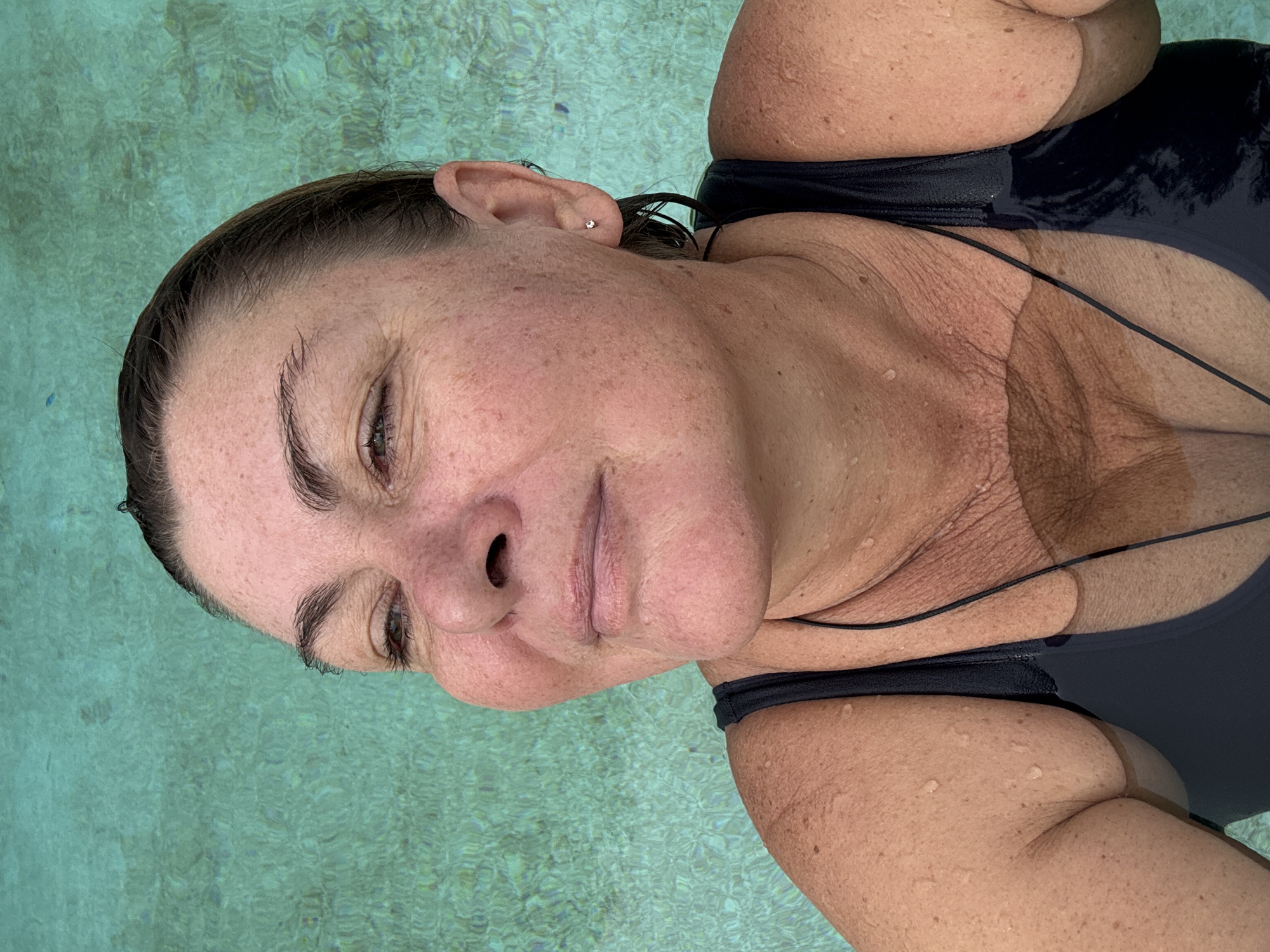 A middle-aged woman with wet, dark brown hair slicked back is in a turquoise body of water, likely a swimming pool, based on the clear, textured surface and the woman's black swimsuit. She is looking to her right with her eyes partially closed, her head tilted slightly upwards. Water droplets are visible on her skin and shoulders. She is wearing a simple black swimsuit with thin straps. The background is entirely the water, which appears to be shallow and has a rippled surface. The lighting suggests it is daytime. The scene is a close-up portrait of the woman.