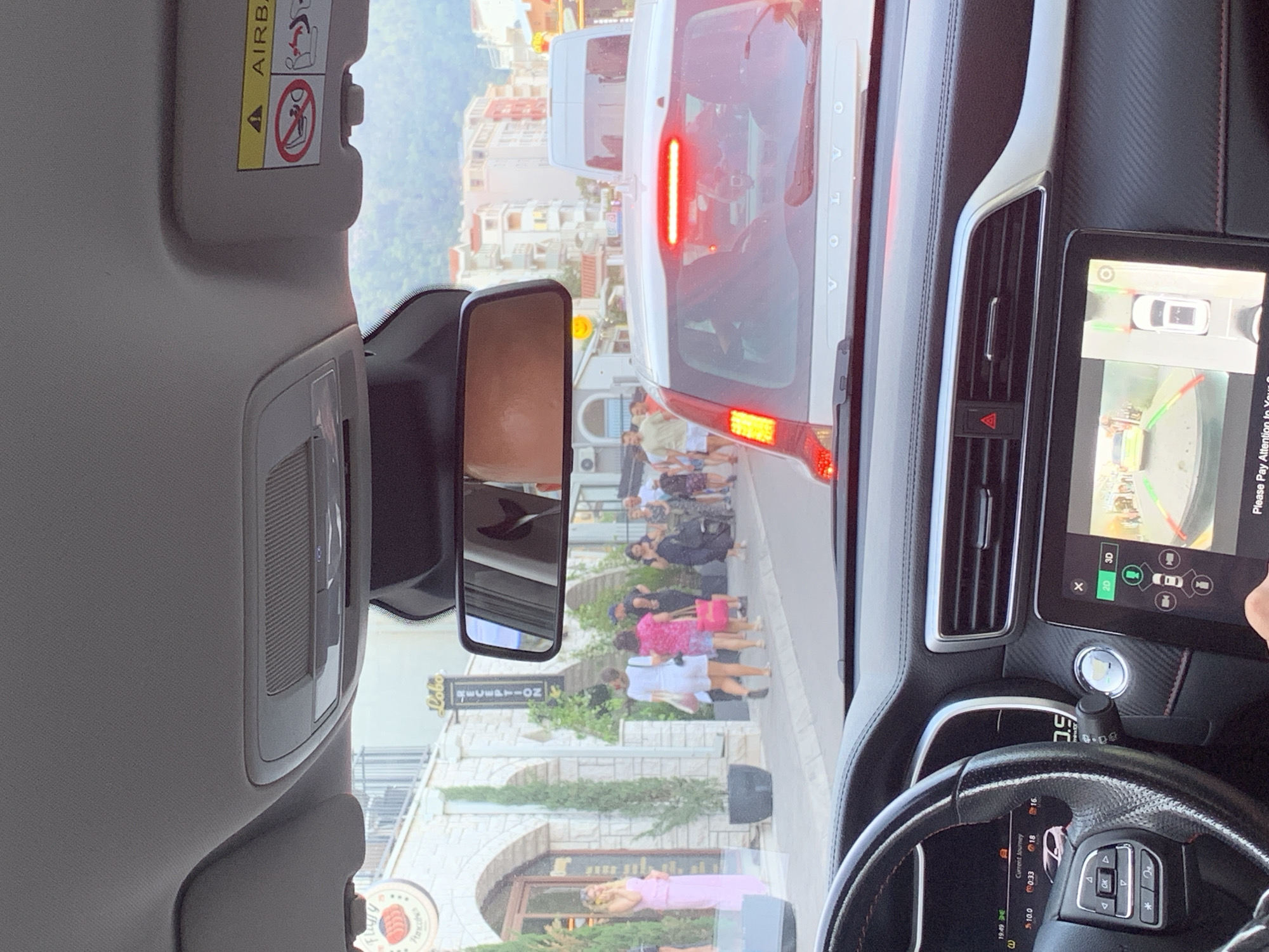The image is taken from the interior of a car in Budva, Montenegro, likely a street in the old town or a similar bustling area. The view is through the windshield and includes the car's dashboard, steering wheel, and rearview mirror.
In the foreground, the car's dashboard is visible, with a steering wheel on the left and a digital display screen on the right showing a rearview camera feed. The rearview camera displays a street scene with other vehicles and pedestrians.
Outside the car, there is a street scene with several people walking on a sidewalk. Buildings line the street, some with shops and signs. There are also trees and potted plants. A white Volvo is visible directly in front of the car, with its brake lights illuminated, indicating traffic congestion. Other vehicles, including a white van, are also present. The architecture of the buildings suggests a Mediterranean or European setting.
The overall impression is a snapshot of everyday life in a busy street in Budva, captured from the perspective of a driver in traffic.