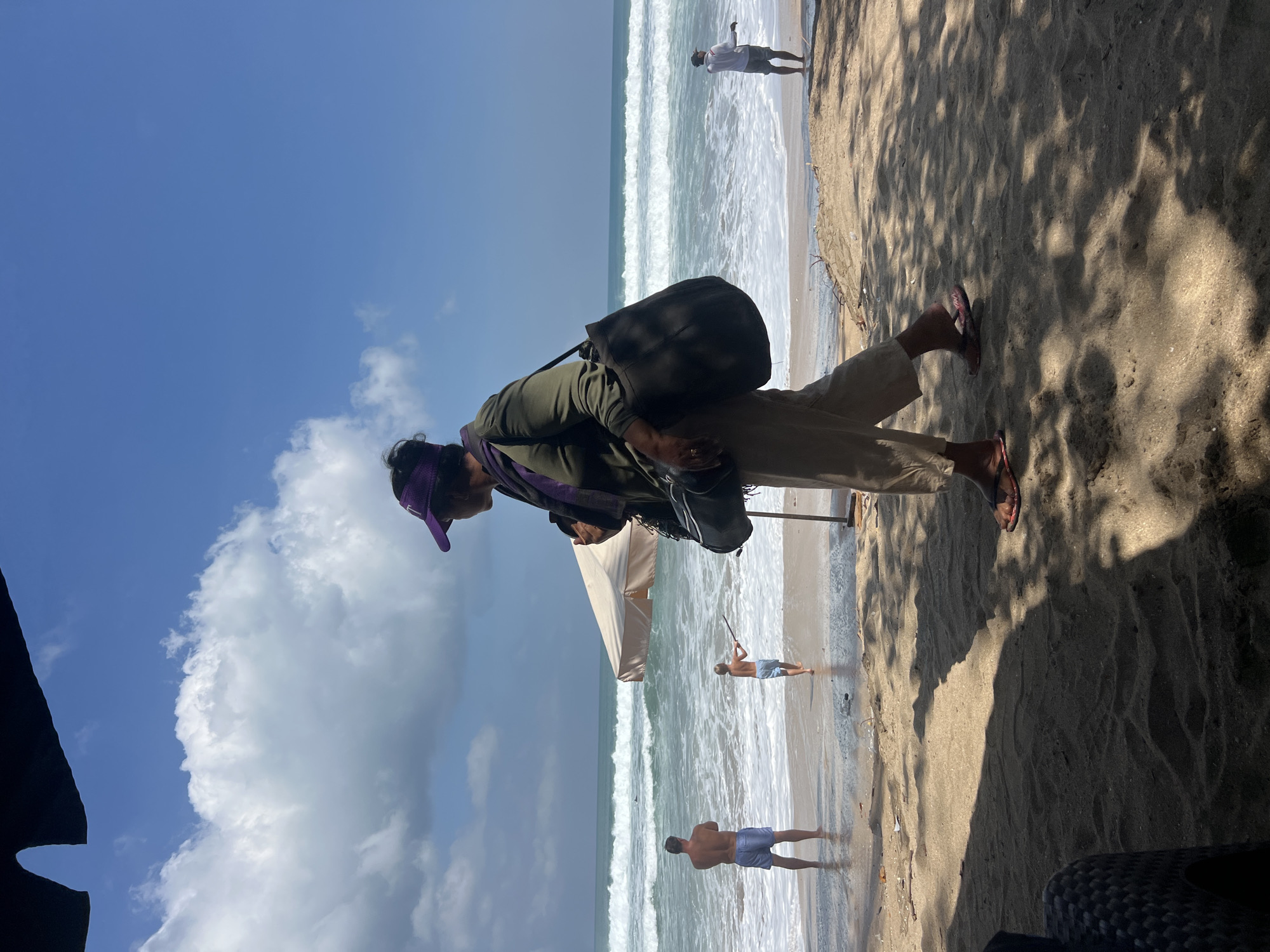 The image depicts a sunny day at Kuta Beach in Kuta, Indonesia, with the ocean and sand being prominent. A woman, viewed from the side and partially obscured by shadow, walks on the sand in the foreground. She is wearing a green long-sleeved shirt, beige pants, a purple visor, and flip-flops. She carries a dark tote bag over her left shoulder and a smaller bag across her body. In the middle ground, the ocean features moderate waves and white surf. Two men are visible near the water's edge. One, shirtless and wearing blue shorts, stands facing the ocean. Another, a child wearing light-colored shorts, is facing away from the camera and holding a stick. A beige beach umbrella stands near them. In the far right background, another person, dressed in a white shirt and dark shorts, stands near the water. The sky is blue with scattered white clouds. The sand in the foreground is textured with sunlight and shadow patterns cast by unseen foliage.